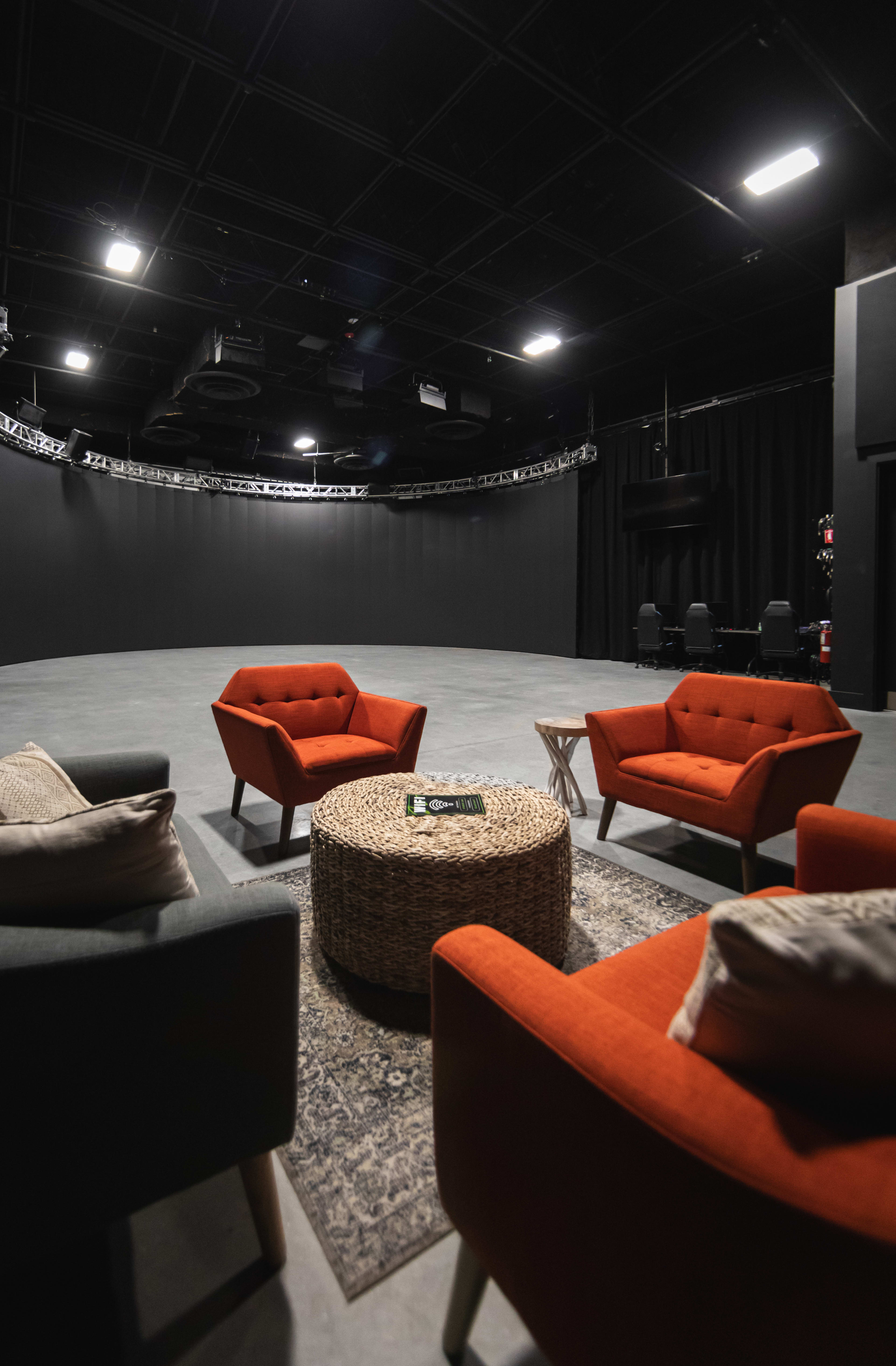 The image shows a spacious, dark studio with gray flooring and a central seating arrangement consisting of two orange armchairs and one gray armchair around a round, woven coffee table.