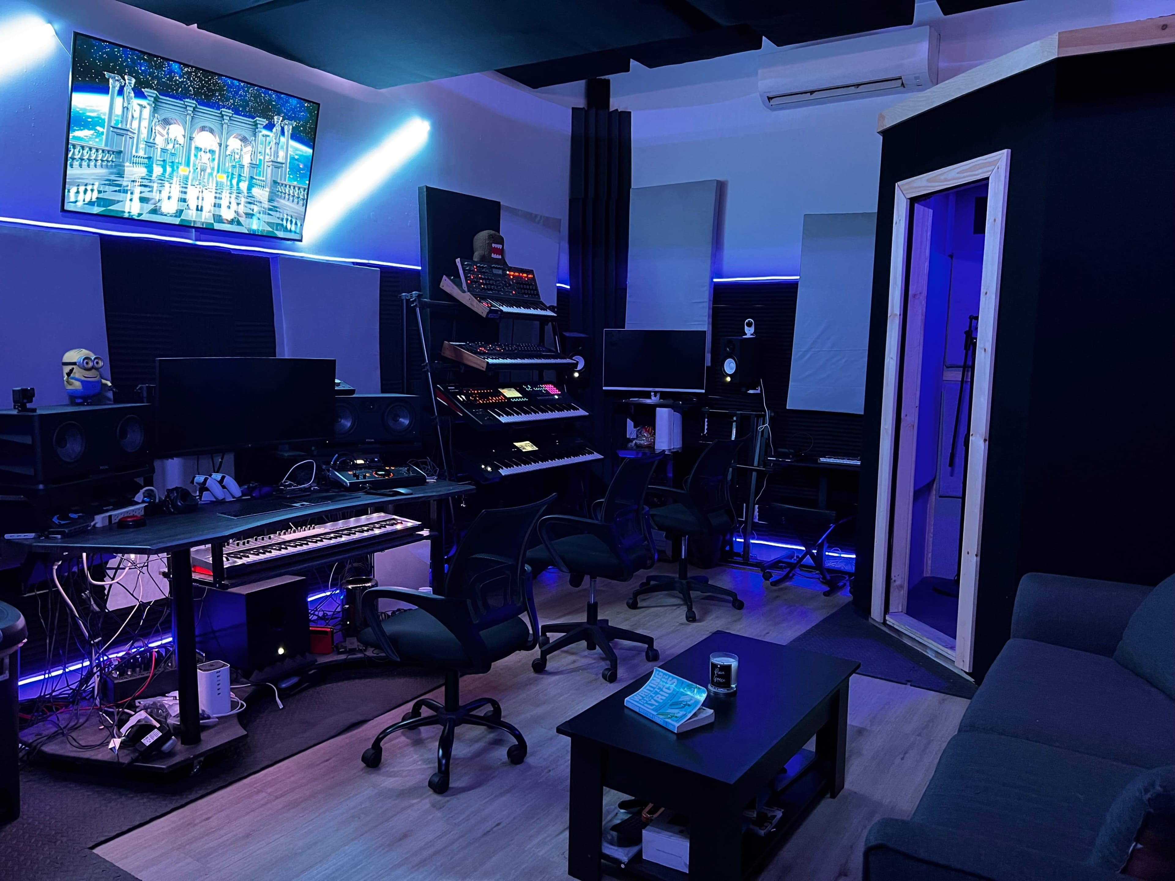 A modern recording studio equipped with multiple computer monitors, musical instruments, and a vocal booth.