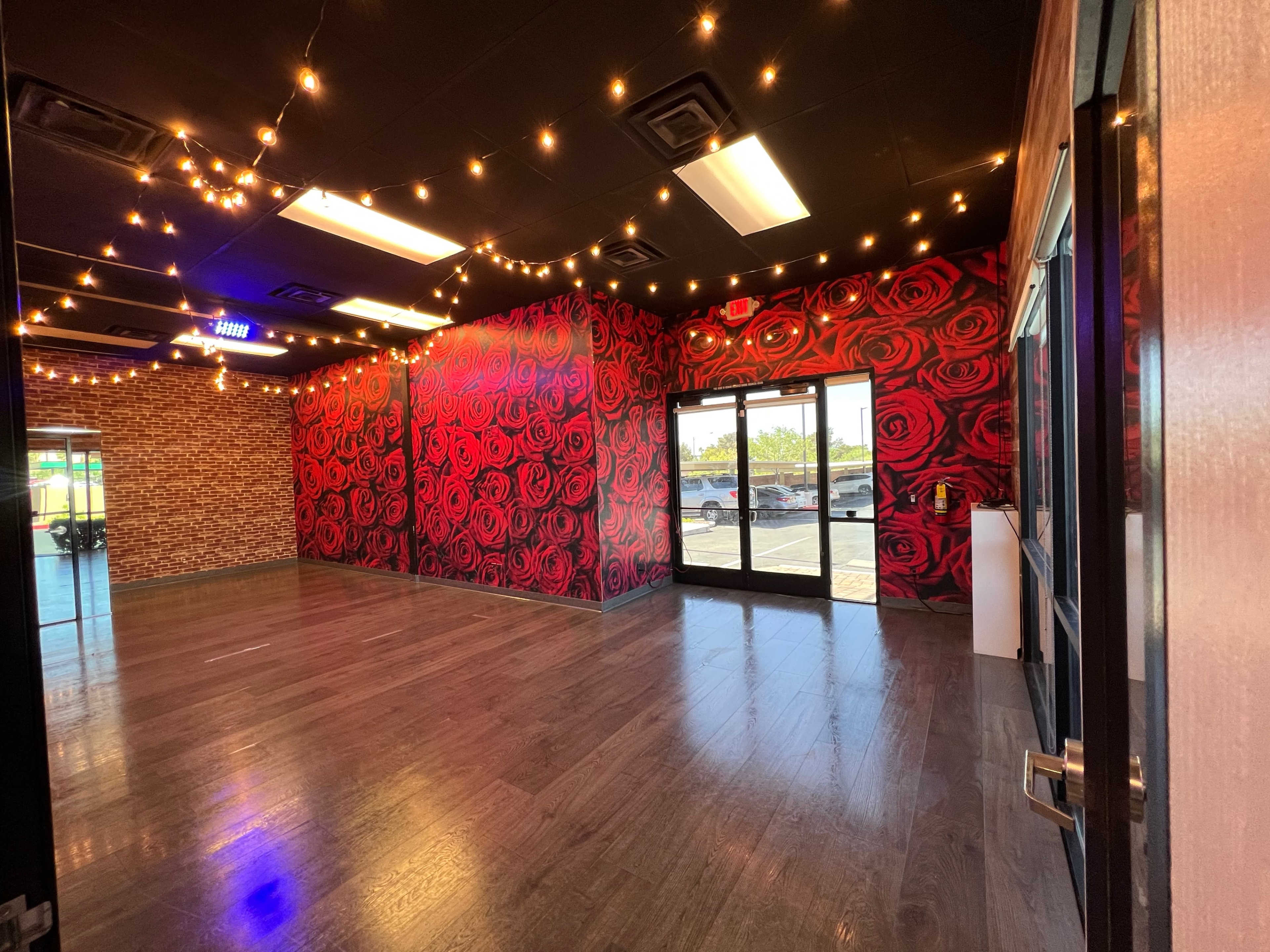 A spacious room with wooden flooring, decorated with a wall featuring large red roses and string lights overhead, leading to glass doors that open to the outside.