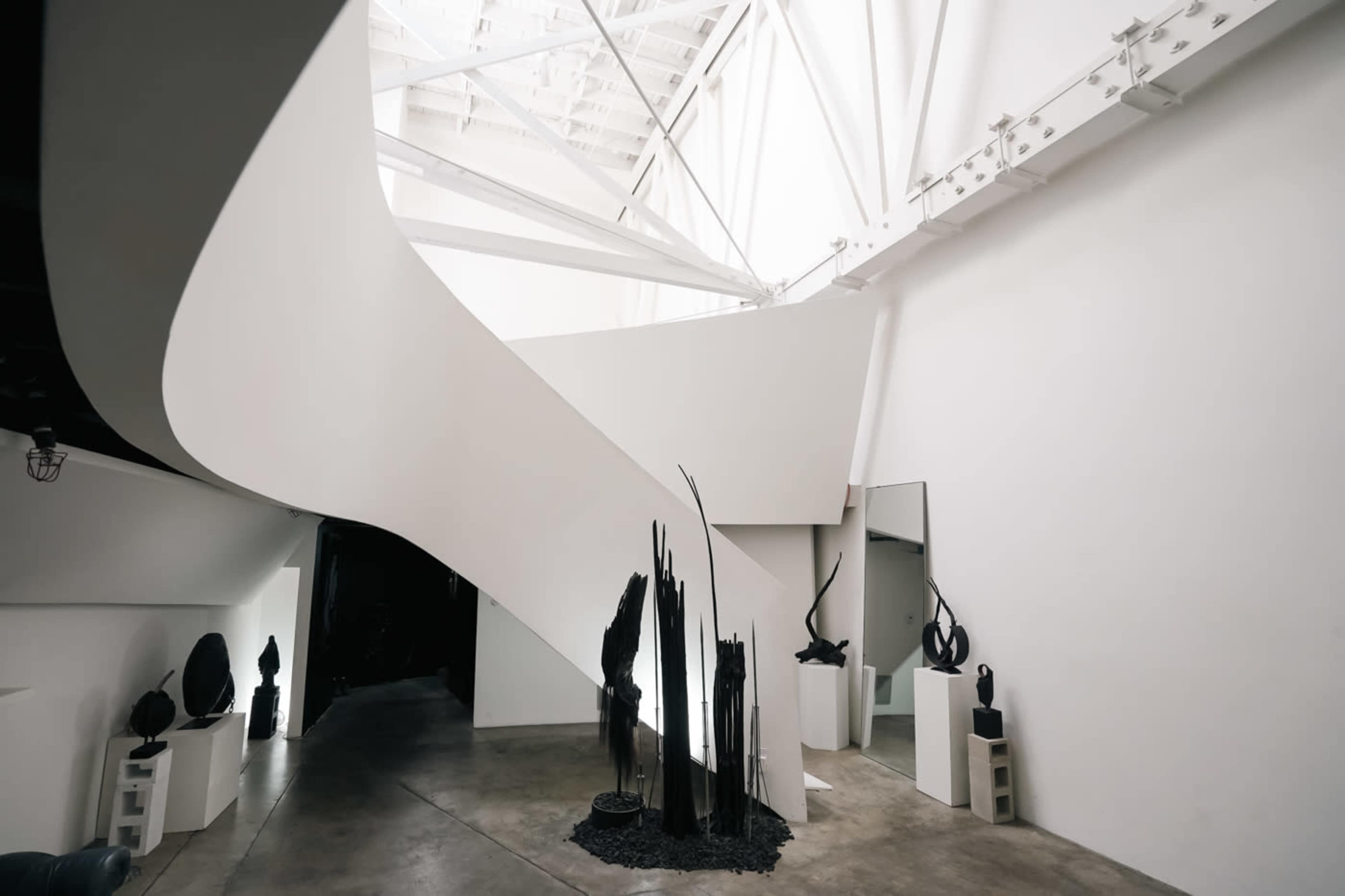 The image shows a modern interior space featuring a spiral staircase, white walls, and various abstract sculptures displayed on pedestals.
