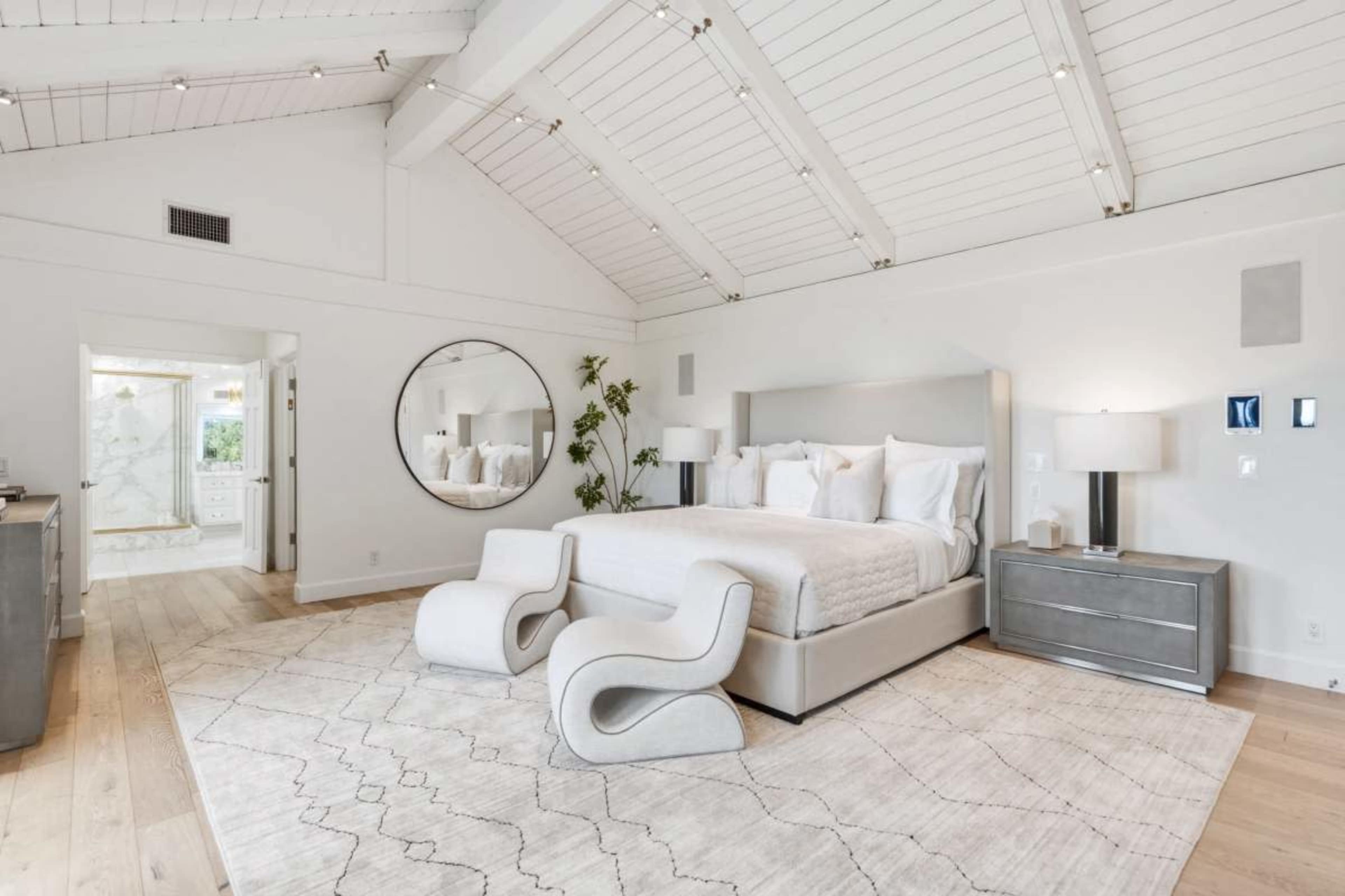 The image shows a spacious, modern bedroom featuring a large bed, geometric chairs, and a circular mirror against a backdrop of high vaulted ceilings.