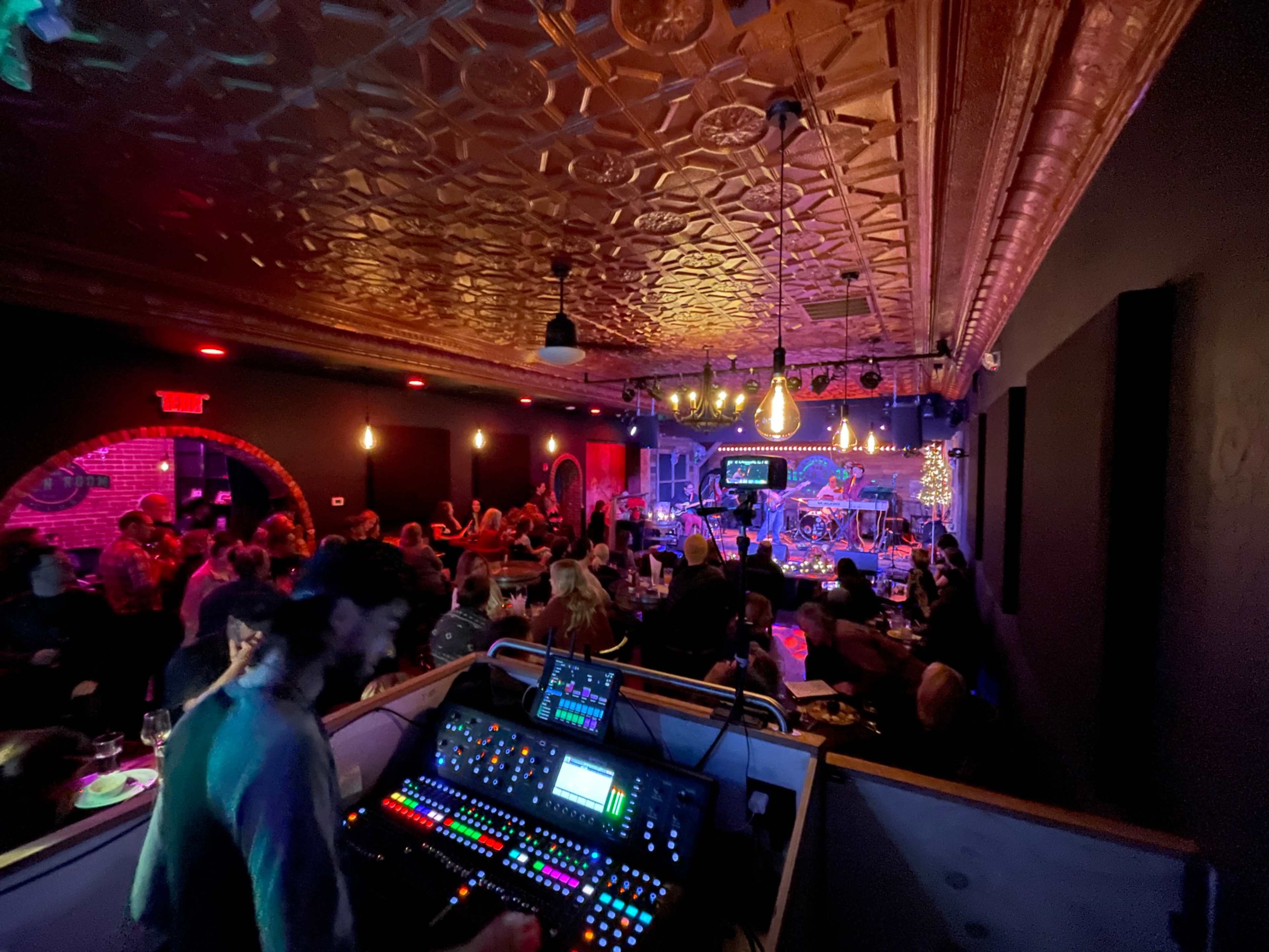 A lively bar scene features a sound mixing console in the foreground and a band performing on stage in the background, with patrons enjoying the atmosphere.