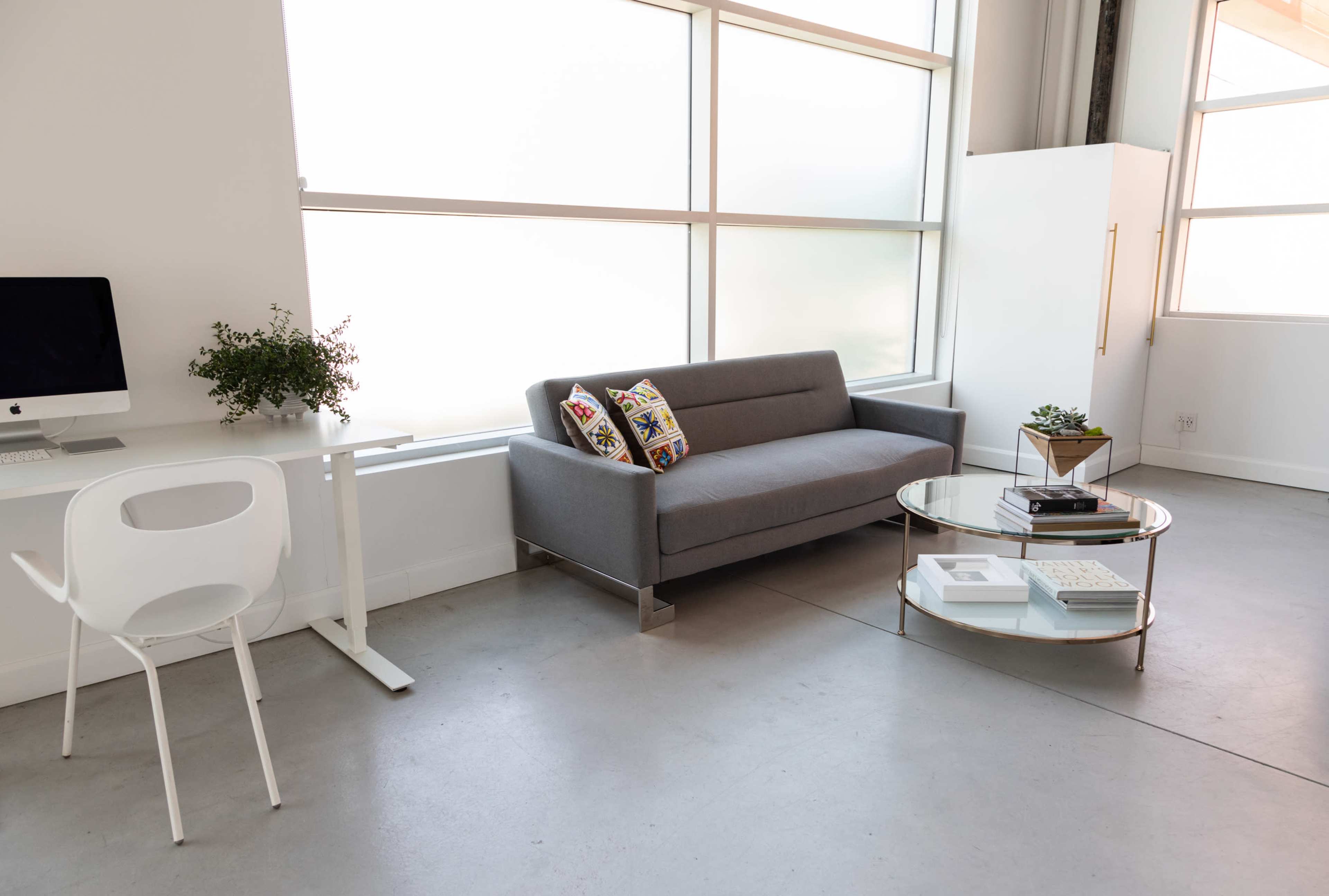 The image shows a modern, minimalist living space featuring a gray sofa, a glass coffee table, a white desk with a computer, and large windows allowing light to enter.