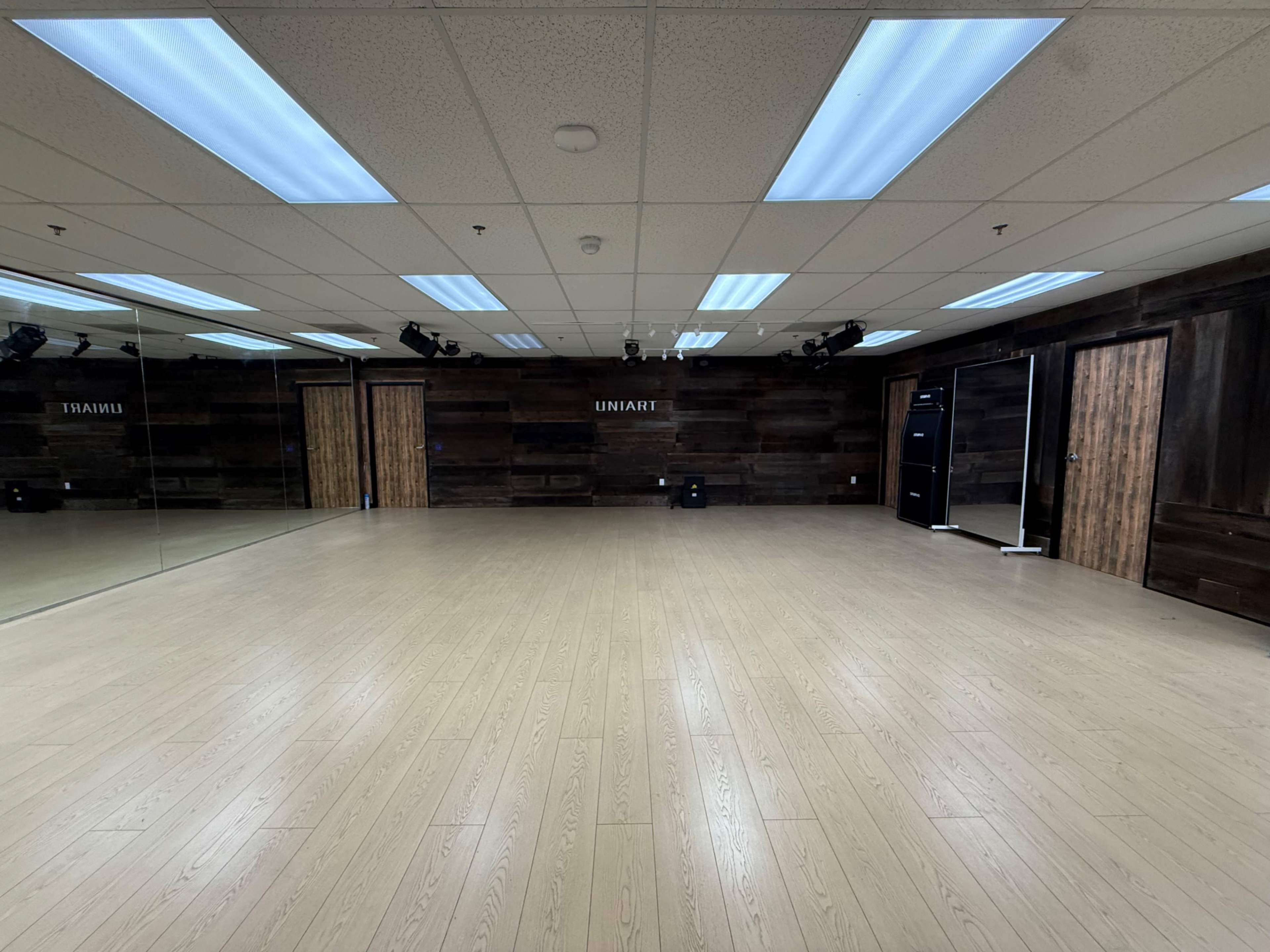 The image shows a spacious, empty dance studio with wooden paneling, hardwood floors, and wall-mounted mirrors.