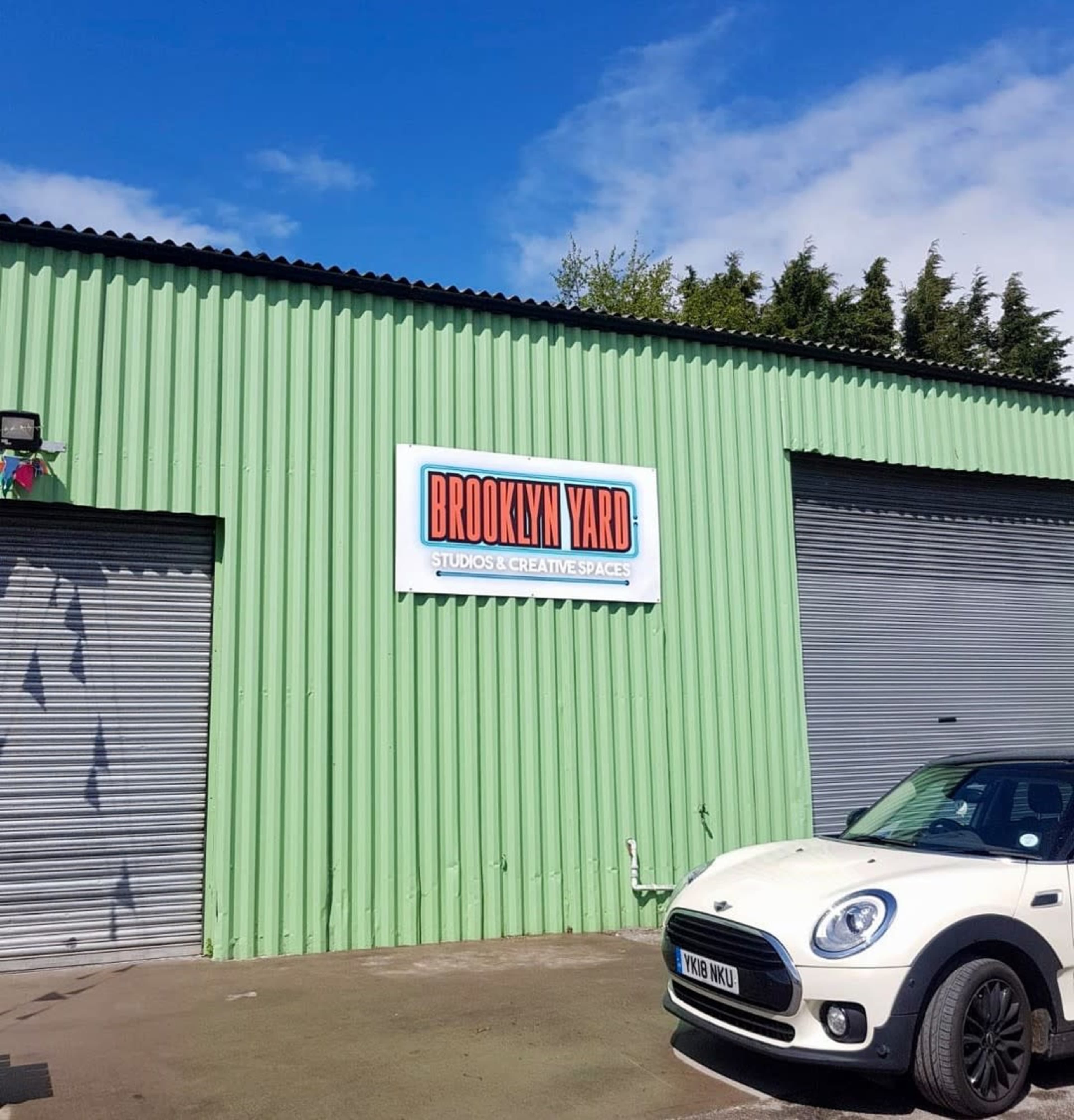A green corrugated metal building with a sign that reads "BROOKLYN YARD STUDIOS & CREATIVE SPACES" and a parked white Mini Cooper in front.