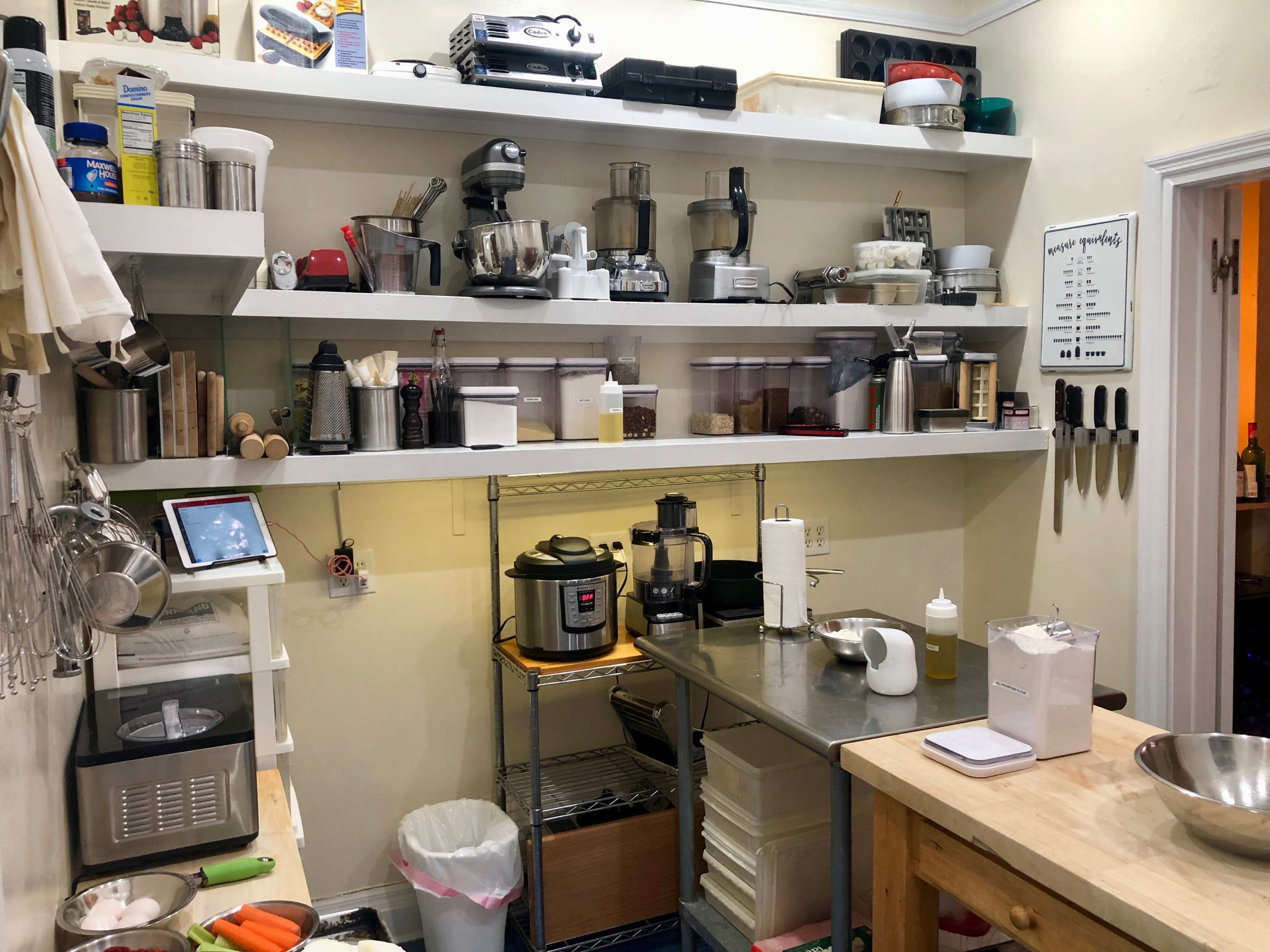 A kitchen workspace features multiple shelves filled with various appliances and utensils, alongside a stainless steel table and garbage bin.