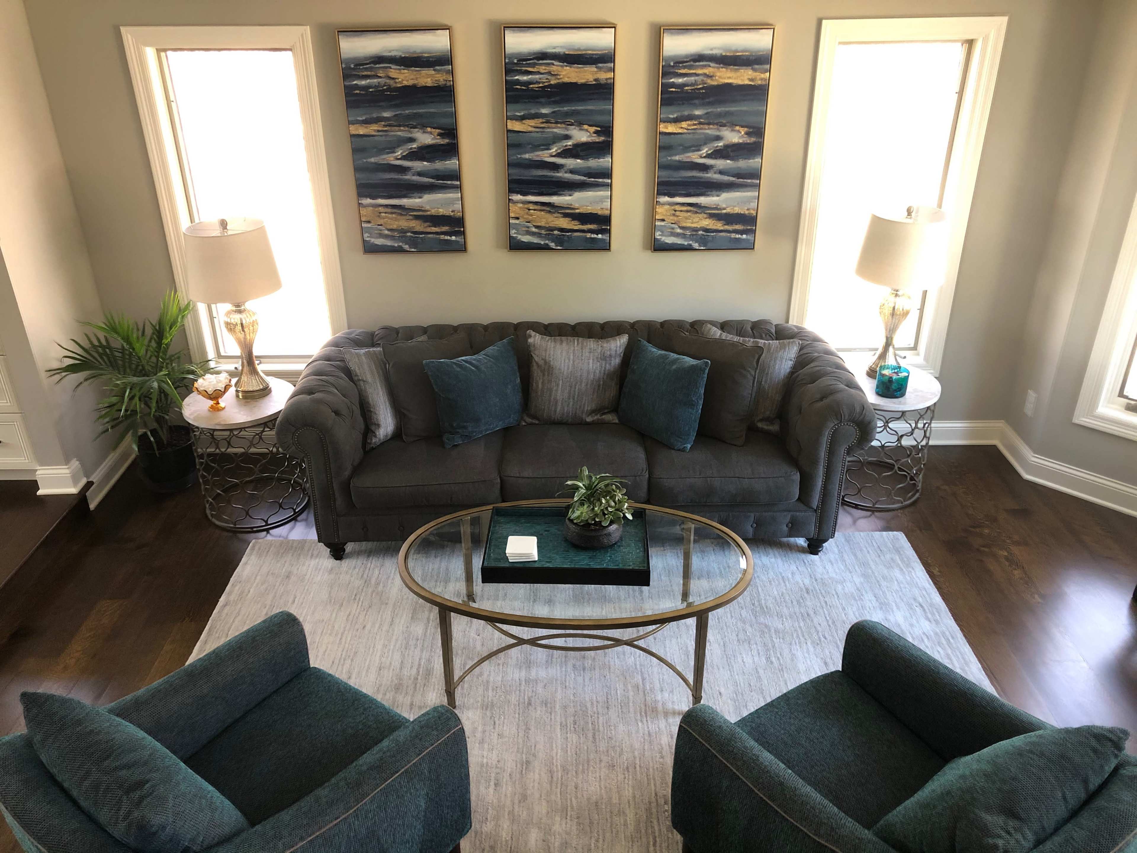 A modern living room with a gray couch, two teal armchairs, a glass coffee table, and three abstract paintings hanging on the wall.