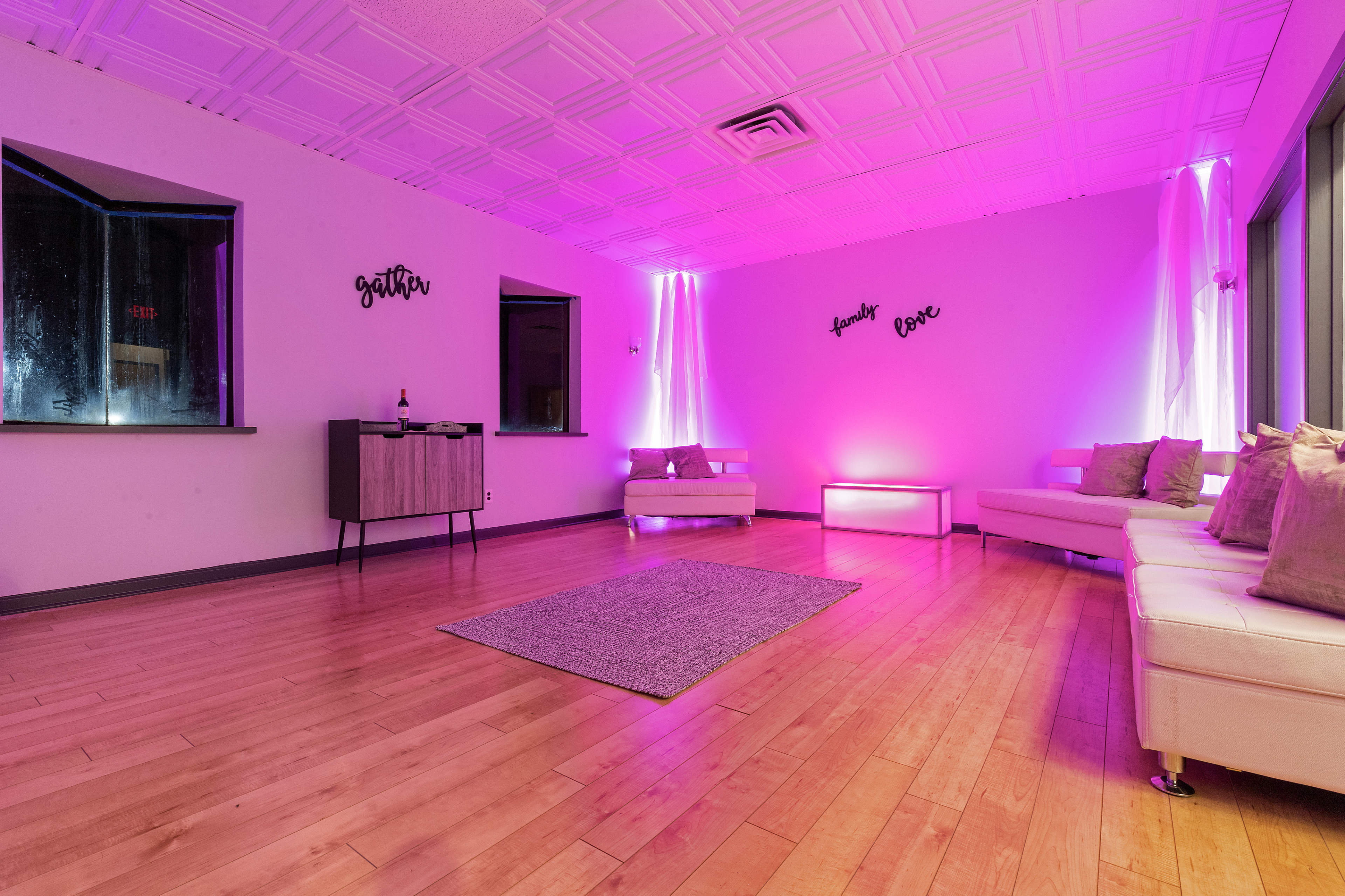 A softly lit lounge features white sofas, a rug, and illuminated walls in purple tones.