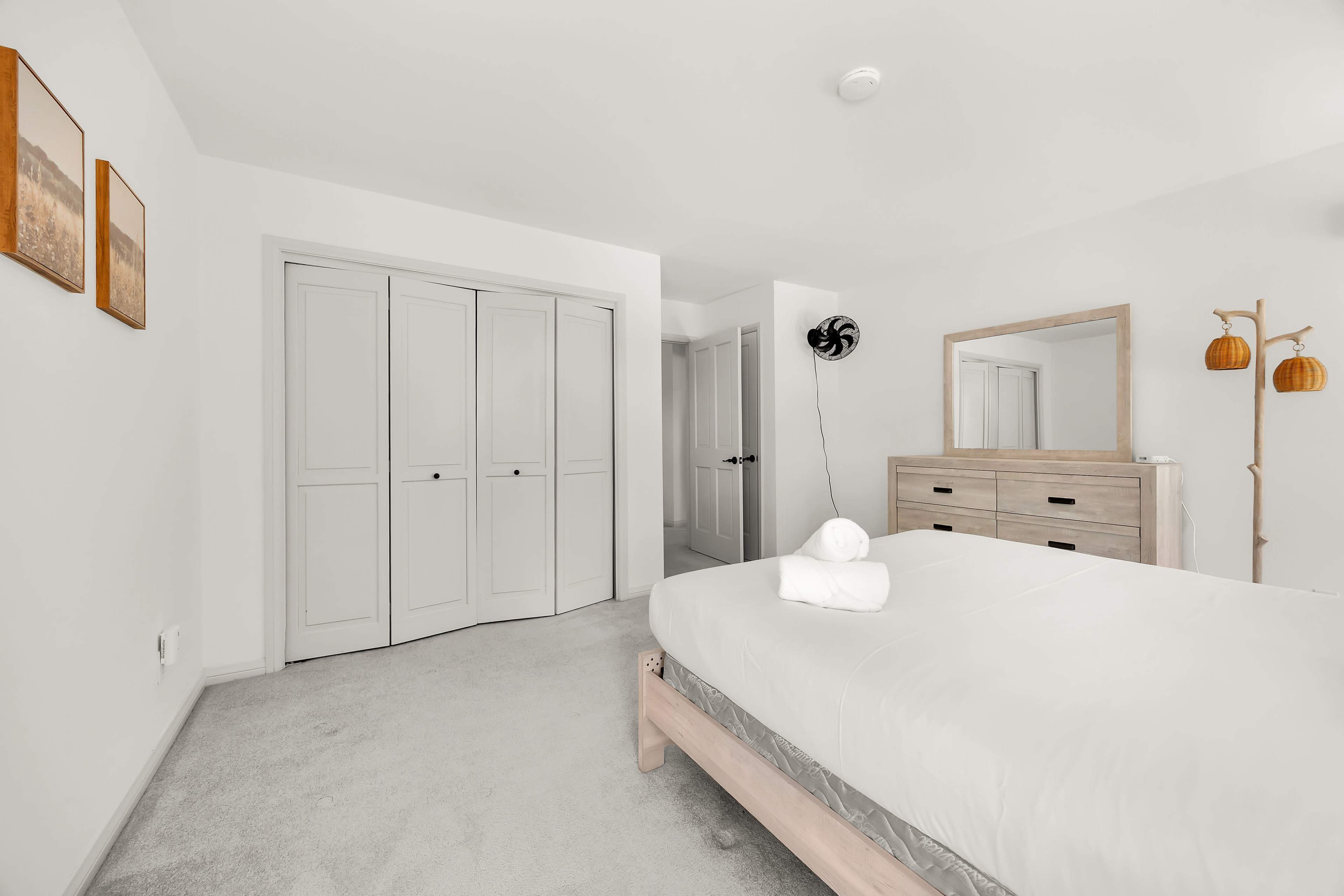 A minimalist bedroom features a bed with a white bedding set, a wooden dresser, a mirror, and a closed closet space with three doors.