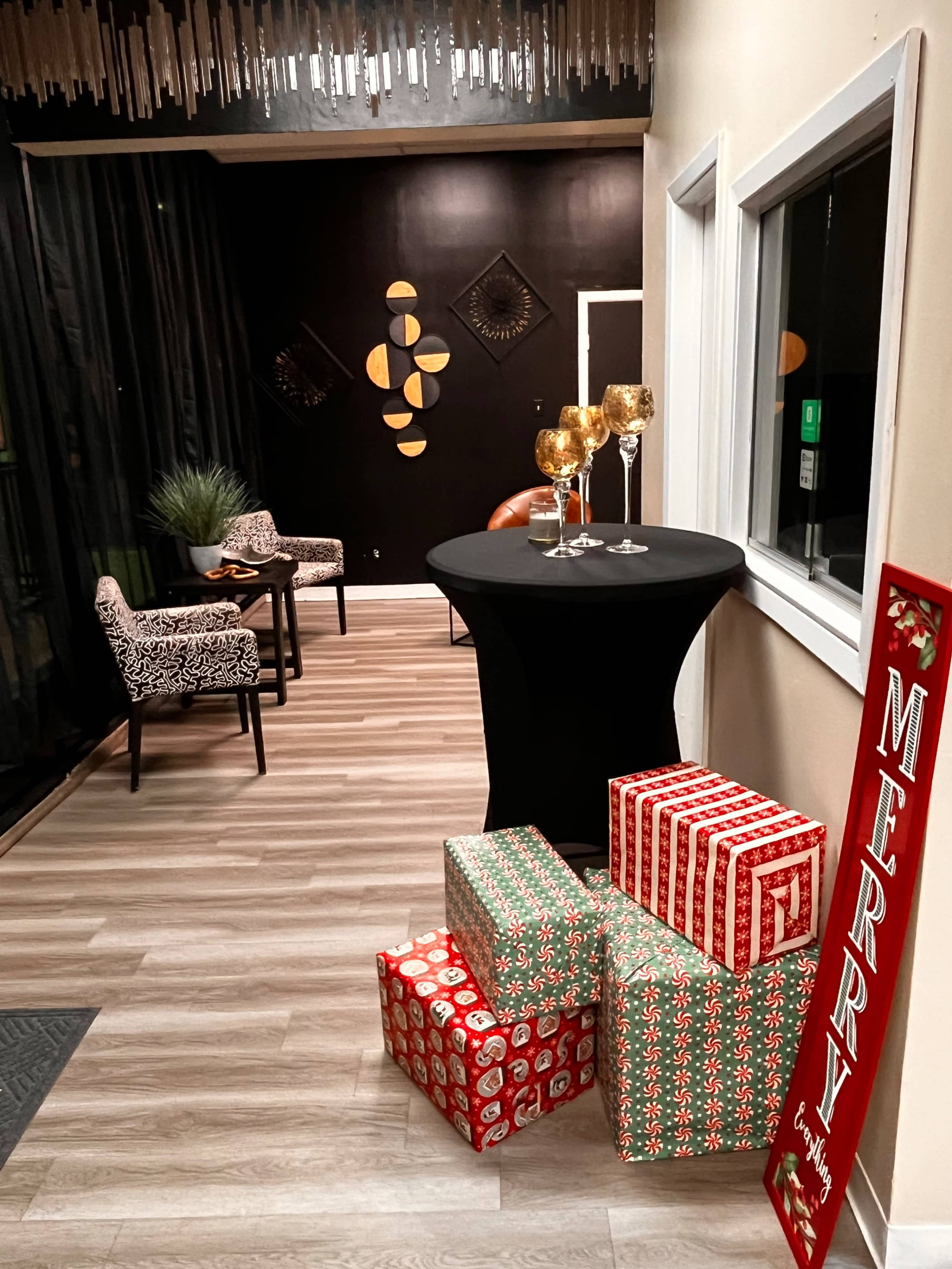 The scene features a decorated hallway with a black table holding three glasses, stylish chairs, and wrapped holiday gifts stacked beside a red sign that says "MERRY."