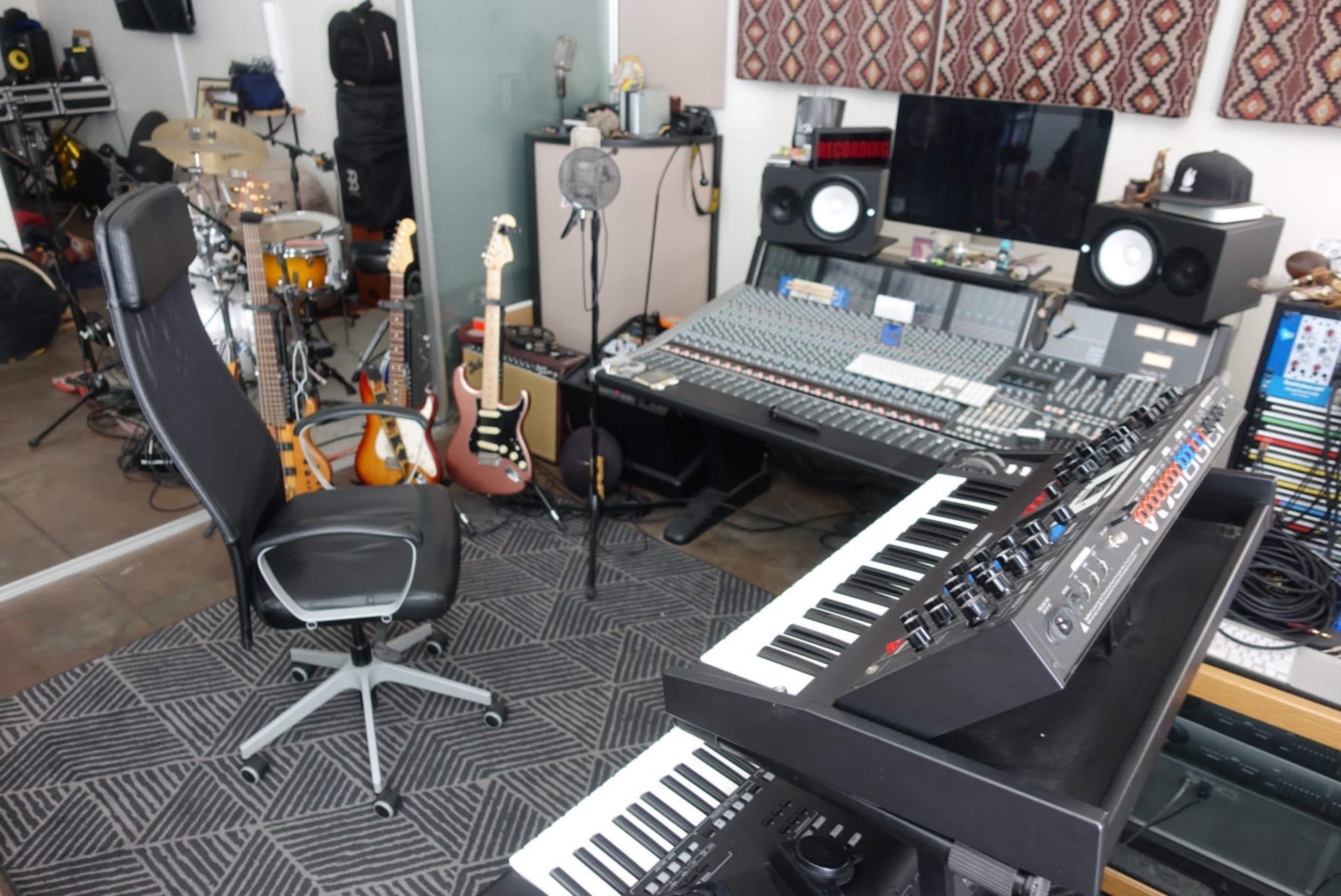 A music studio featuring a mixer, keyboards, guitars, and drums, along with an office chair.