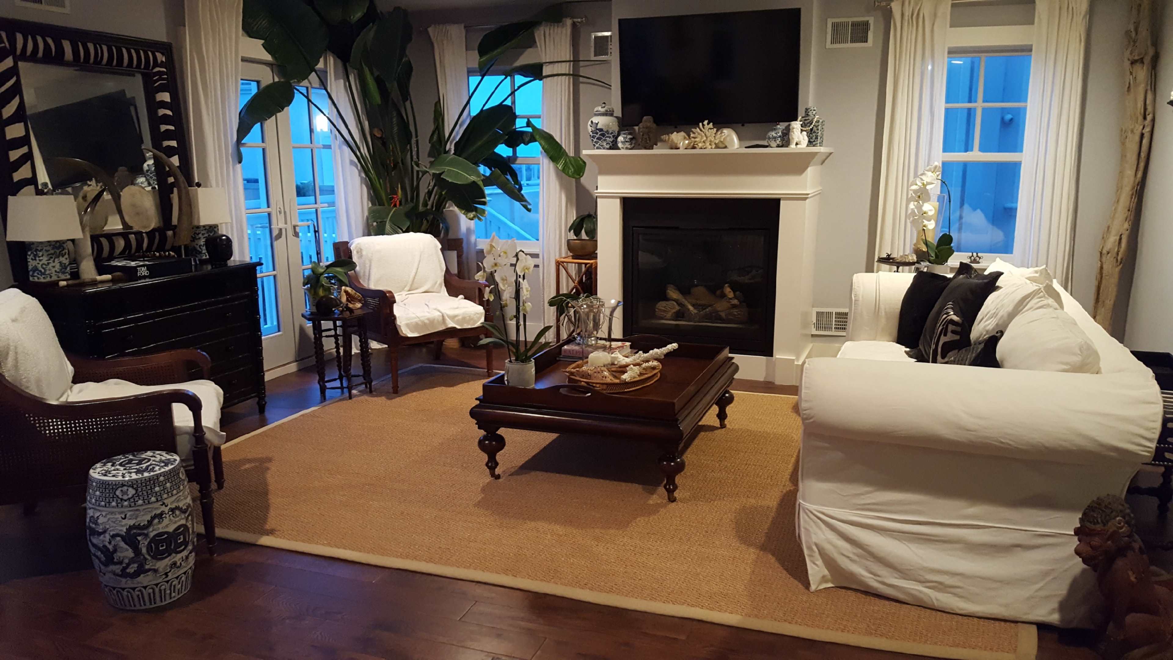 The living room features a large rug, a fireplace, two armchairs, a white sofa, and decorative plants near large windows.