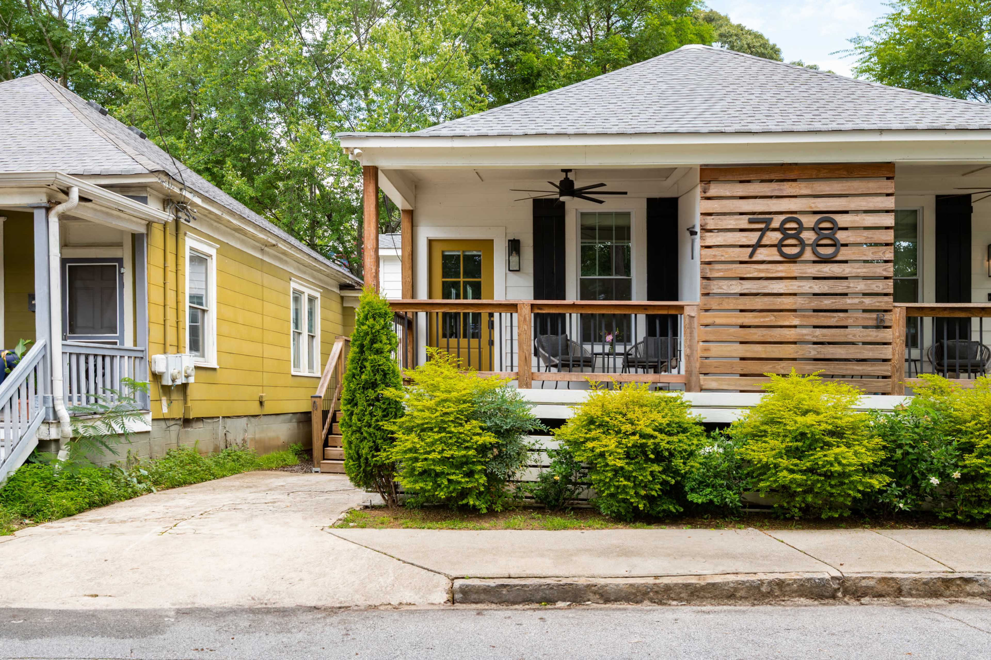 The image shows two houses side by side, one with a yellow exterior and the other featuring a wooden facade, both set on a tree-lined street.