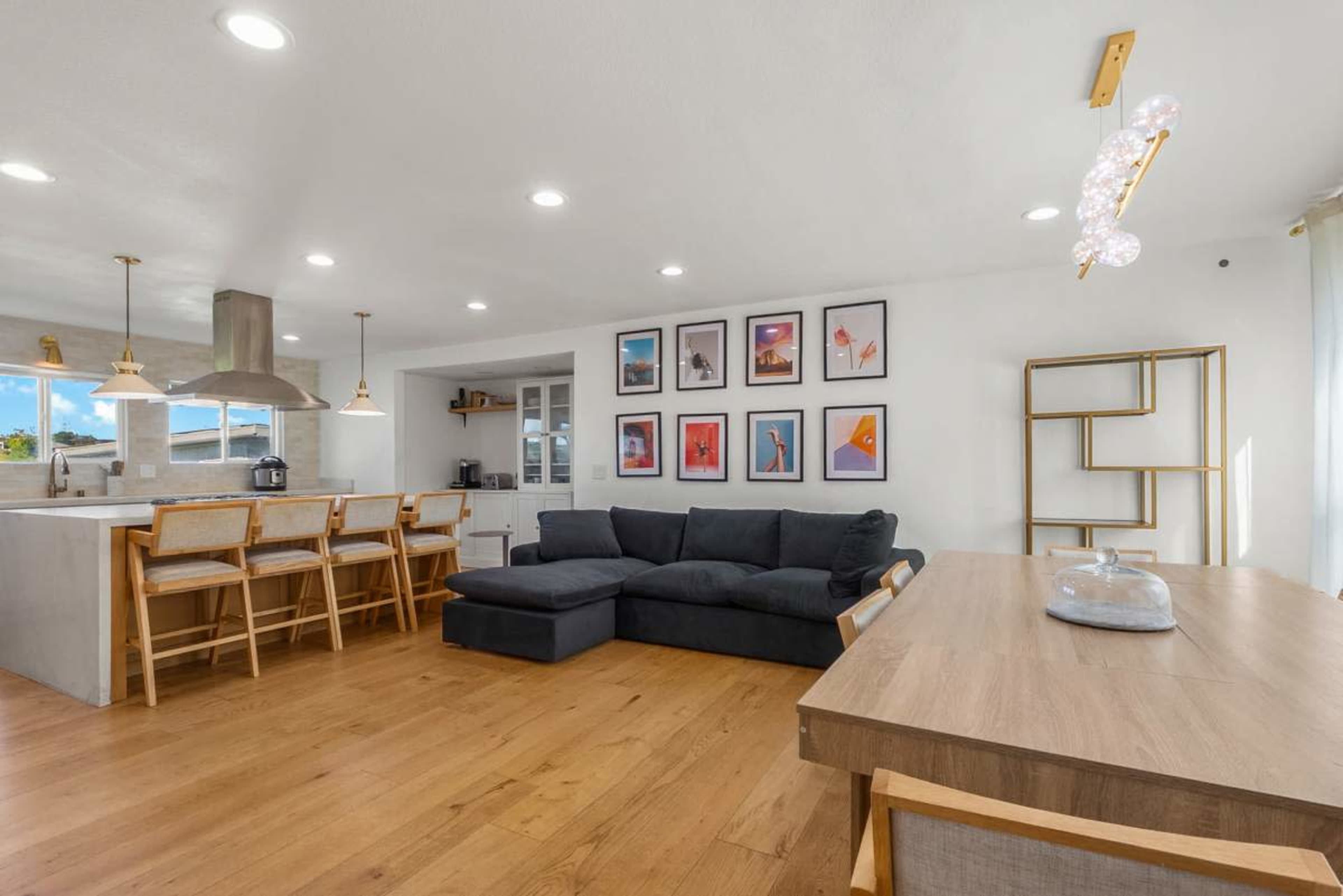 The image shows a modern open-concept living space featuring a sectional sofa, wooden dining table, and multiple framed artworks on the wall.