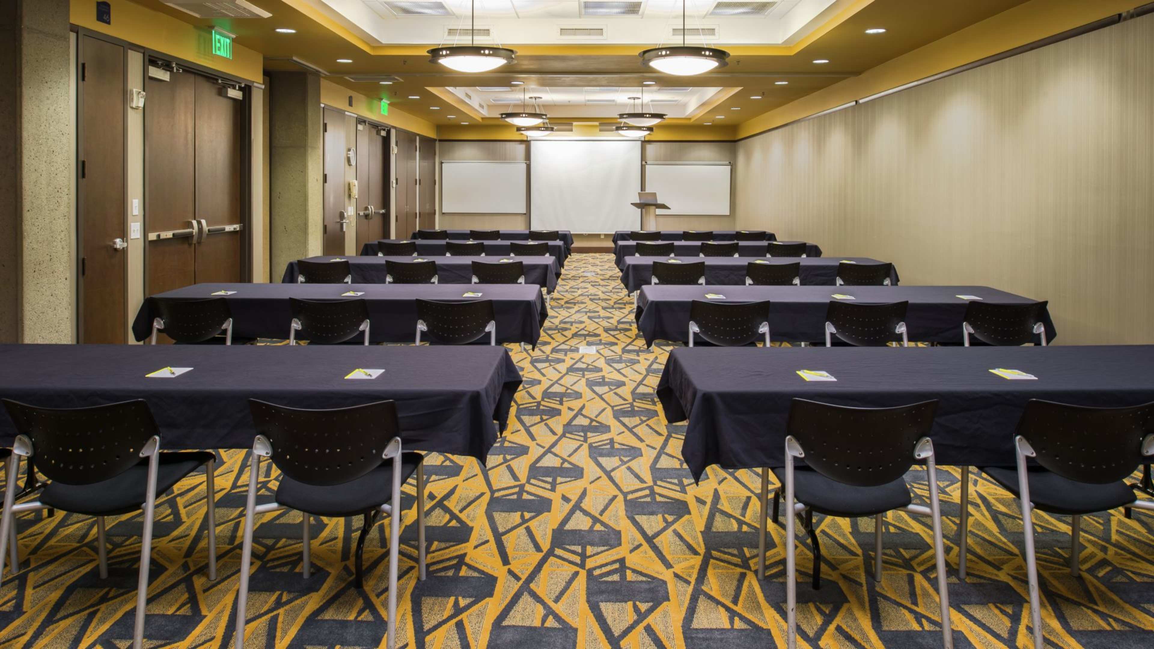 A conference room is arranged with rows of tables and chairs, facing a projector screen and podium against the back wall.