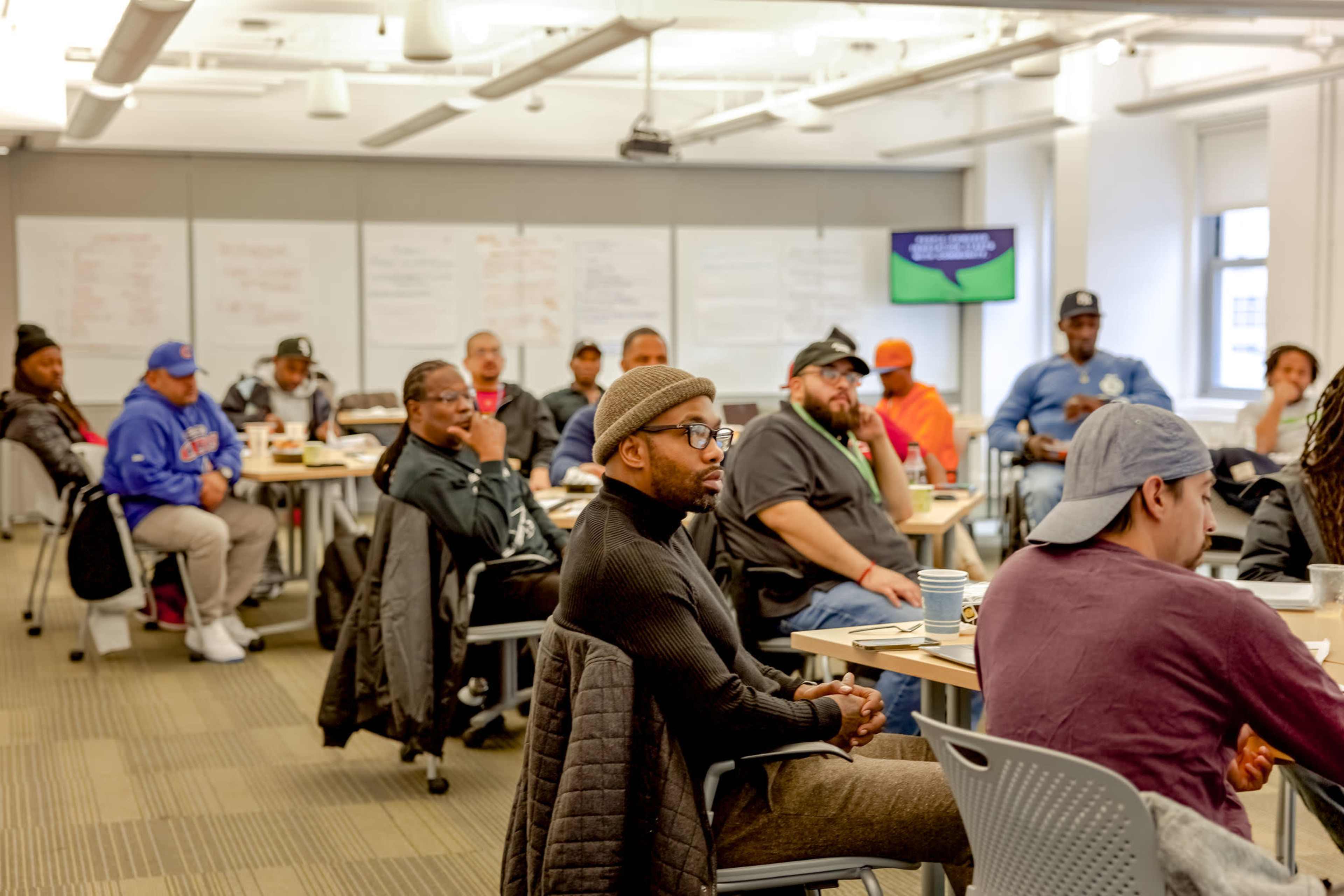 A diverse group of people is seated in a classroom setting, engaged in a discussion, with whiteboards and a presentation screen visible in the background.