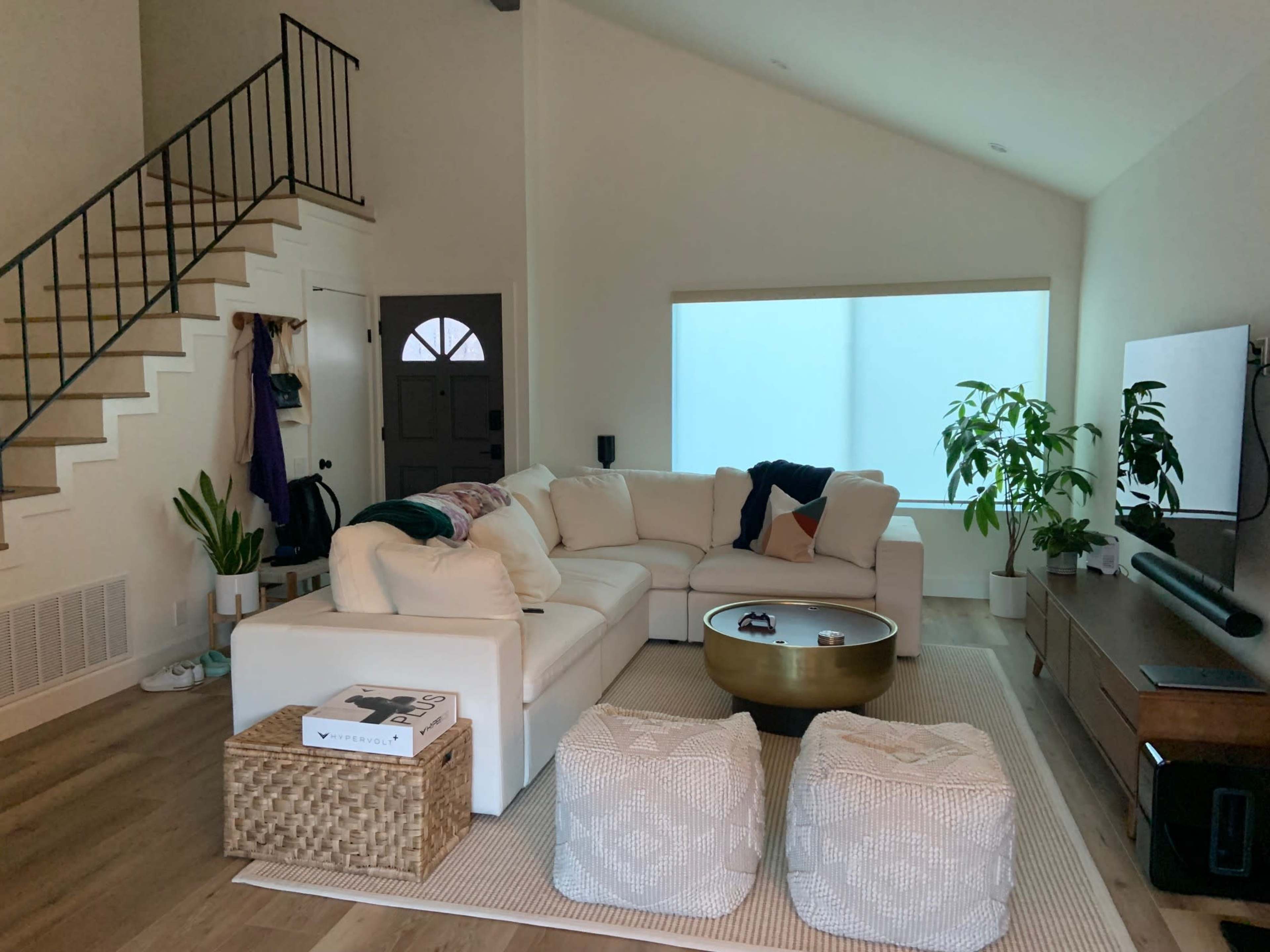 A light-colored sectional sofa is arranged in a living room with a staircase, a large window covered by sheer curtains, and decorative plants.
