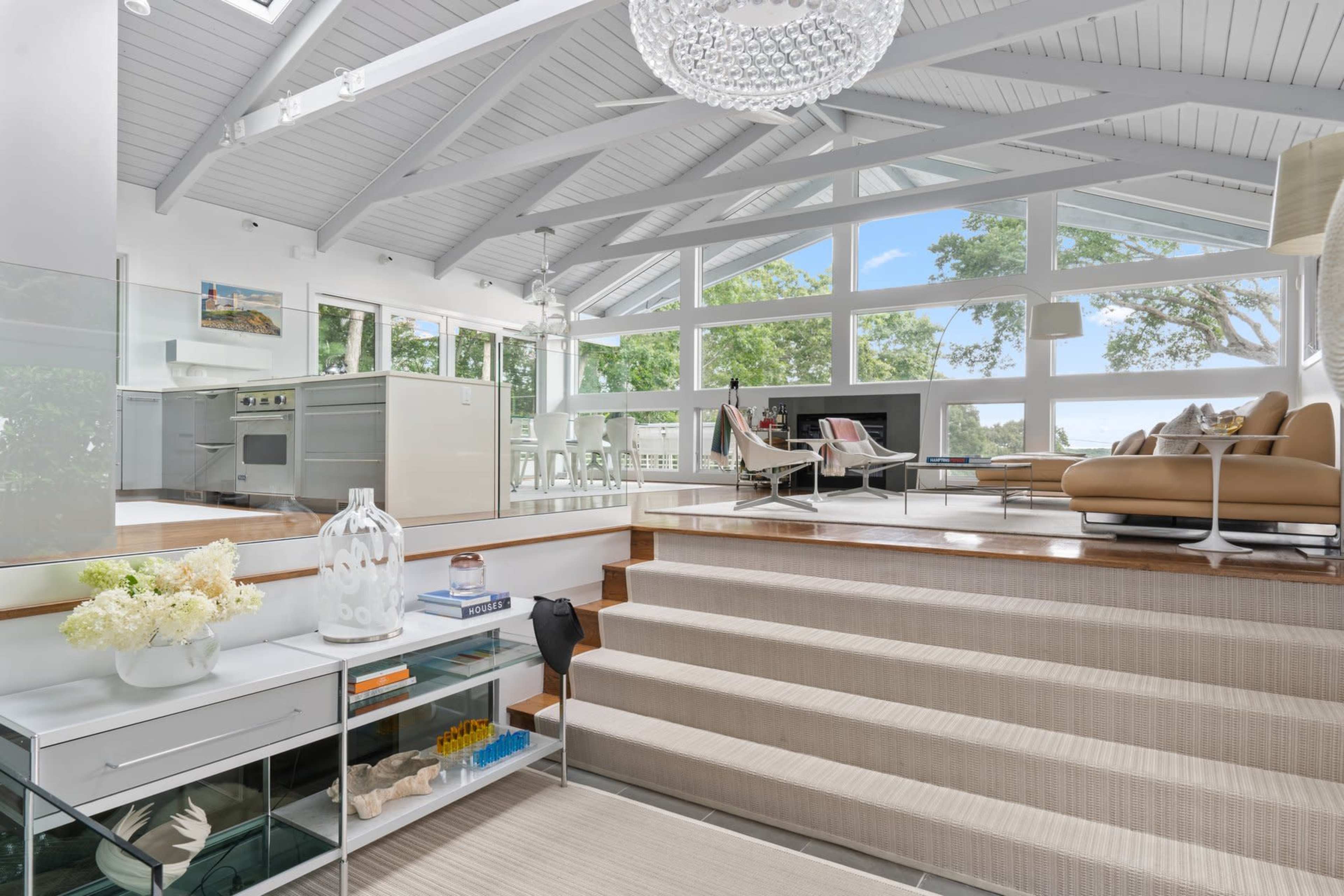 A modern living space with a high ceiling, large windows, and a staircase leading to a dining area and kitchen.