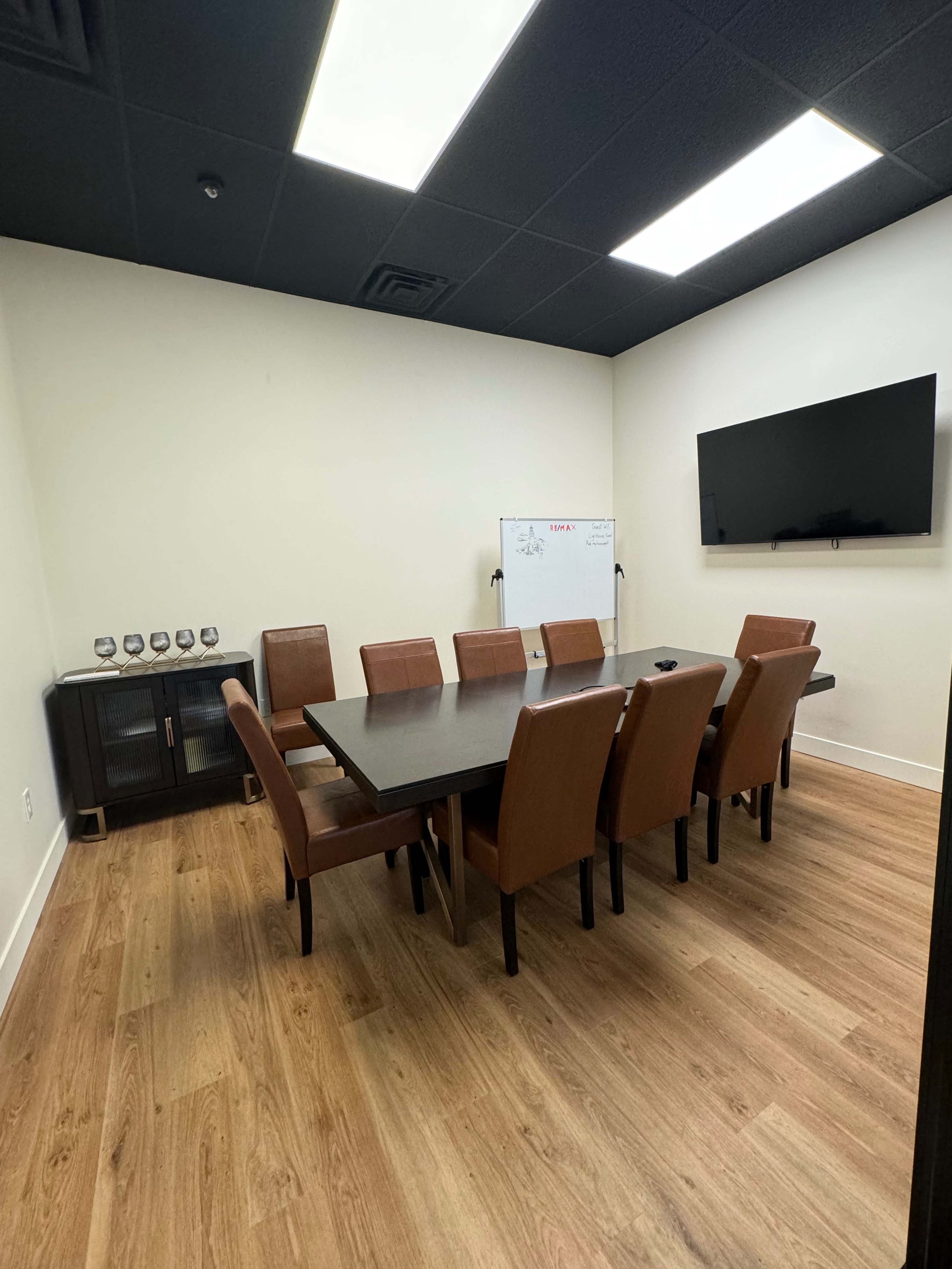 A modern conference room with a large rectangular table surrounded by eight brown chairs, a wall-mounted television, and a whiteboard in the corner.