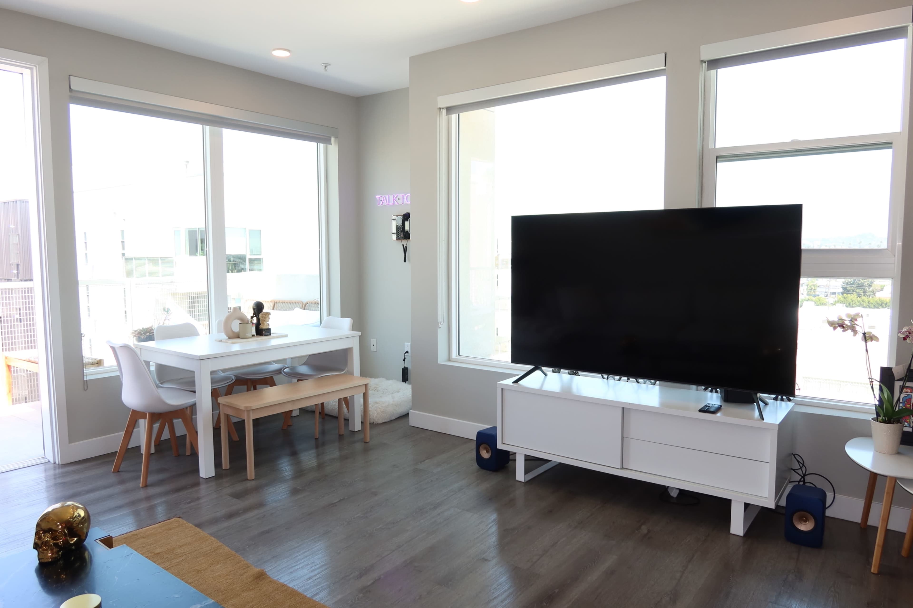 The image shows a modern living room with a large television, a white dining table with chairs, and floor-to-ceiling windows allowing natural light.