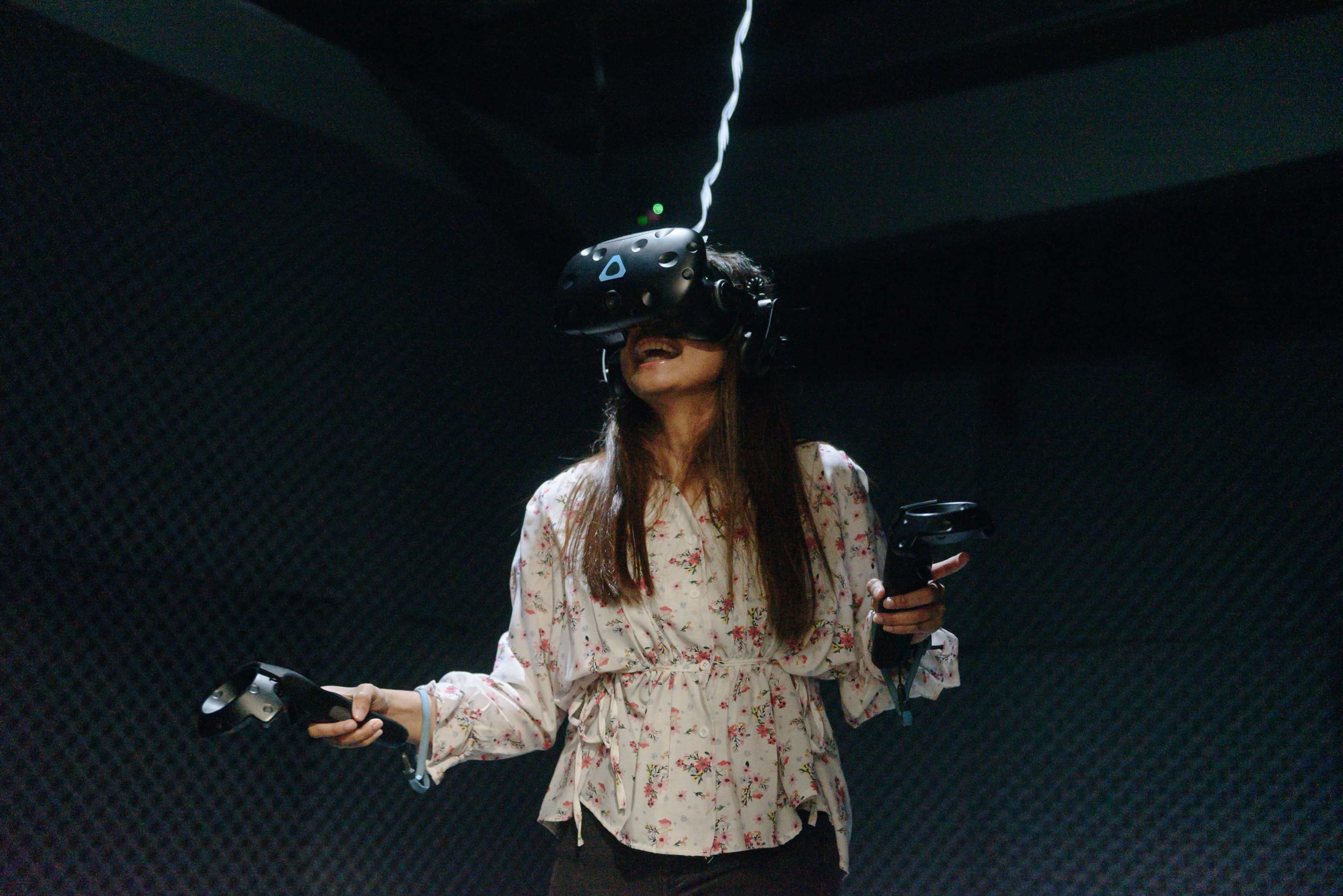 A woman wearing a virtual reality headset holds controllers while standing in a dimly lit room.