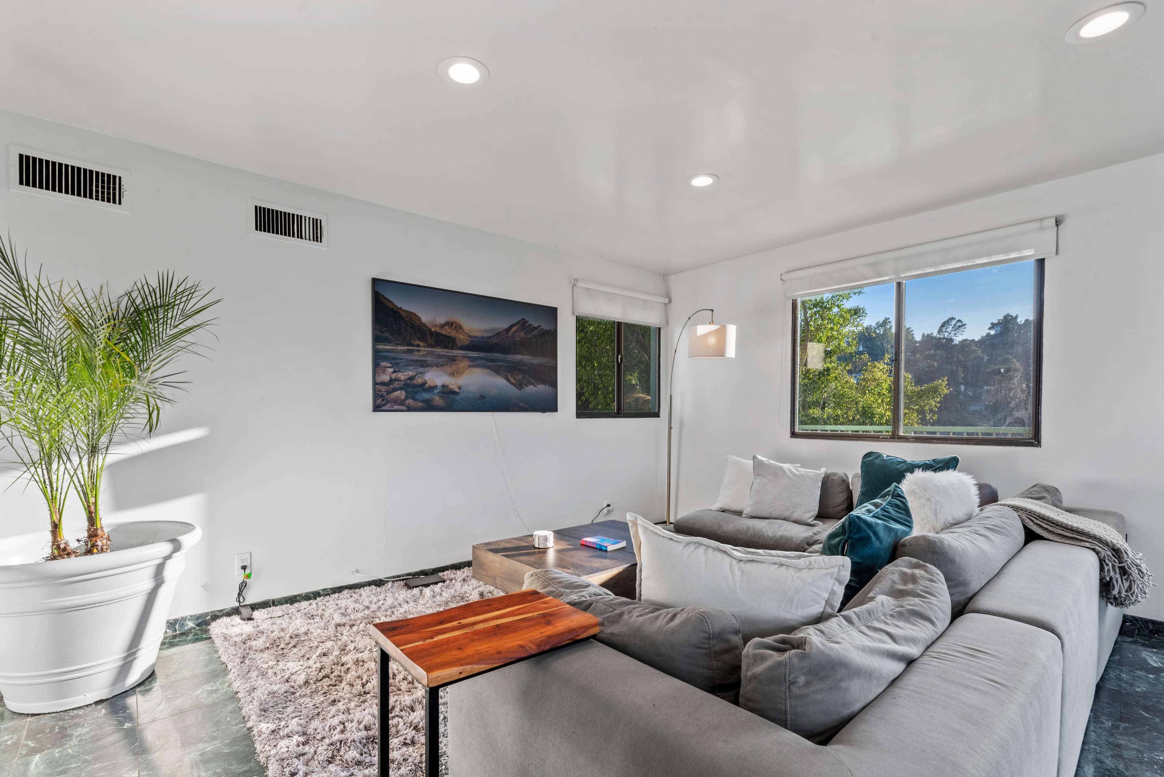 The image shows a modern living room featuring a large sectional sofa, a coffee table, a potted plant, and a scenic landscape painting on the wall, with natural light coming through the windows.