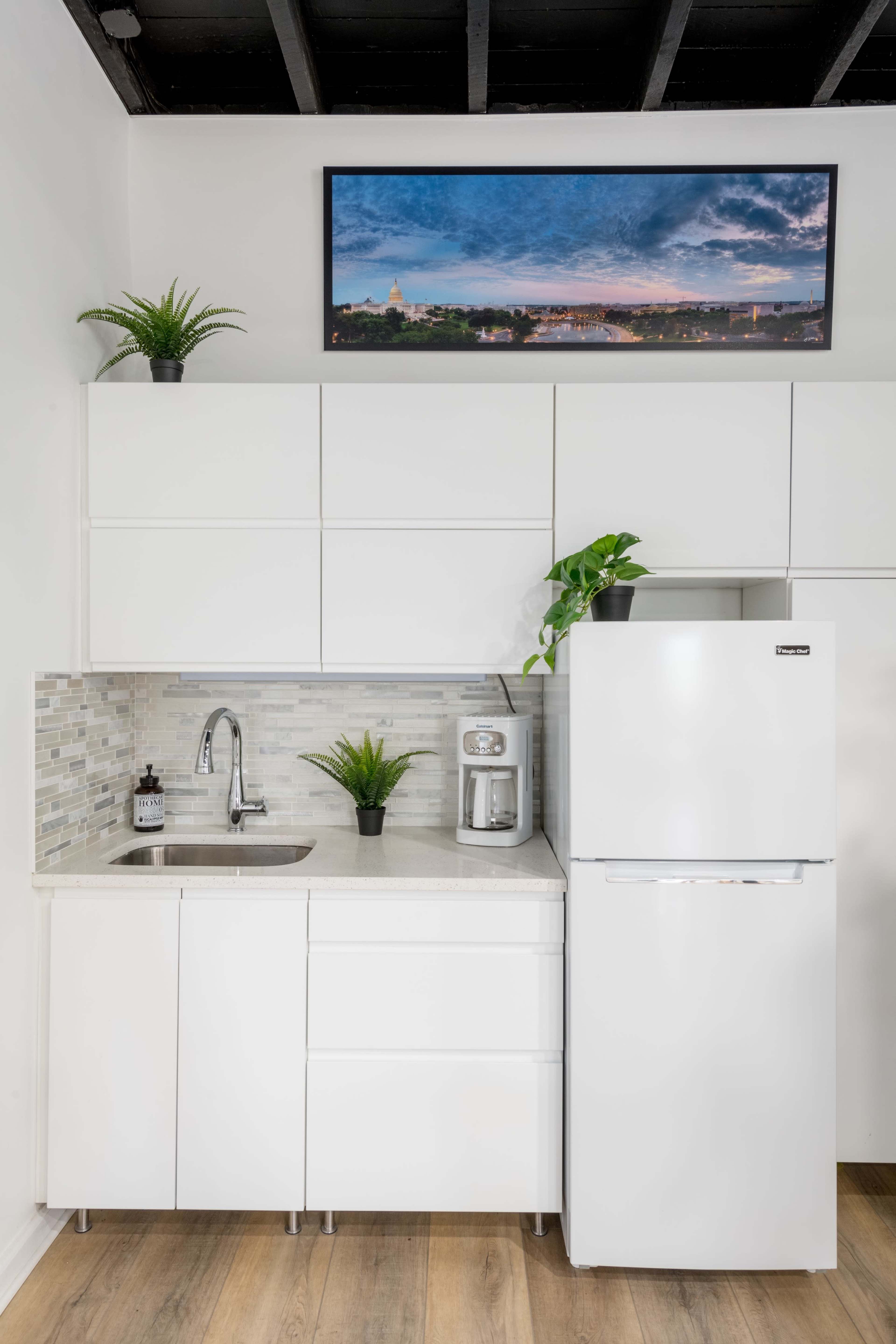 A modern kitchen setup with white cabinets, a sink, a coffee maker, and a white refrigerator, under a framed photo of a landscape.