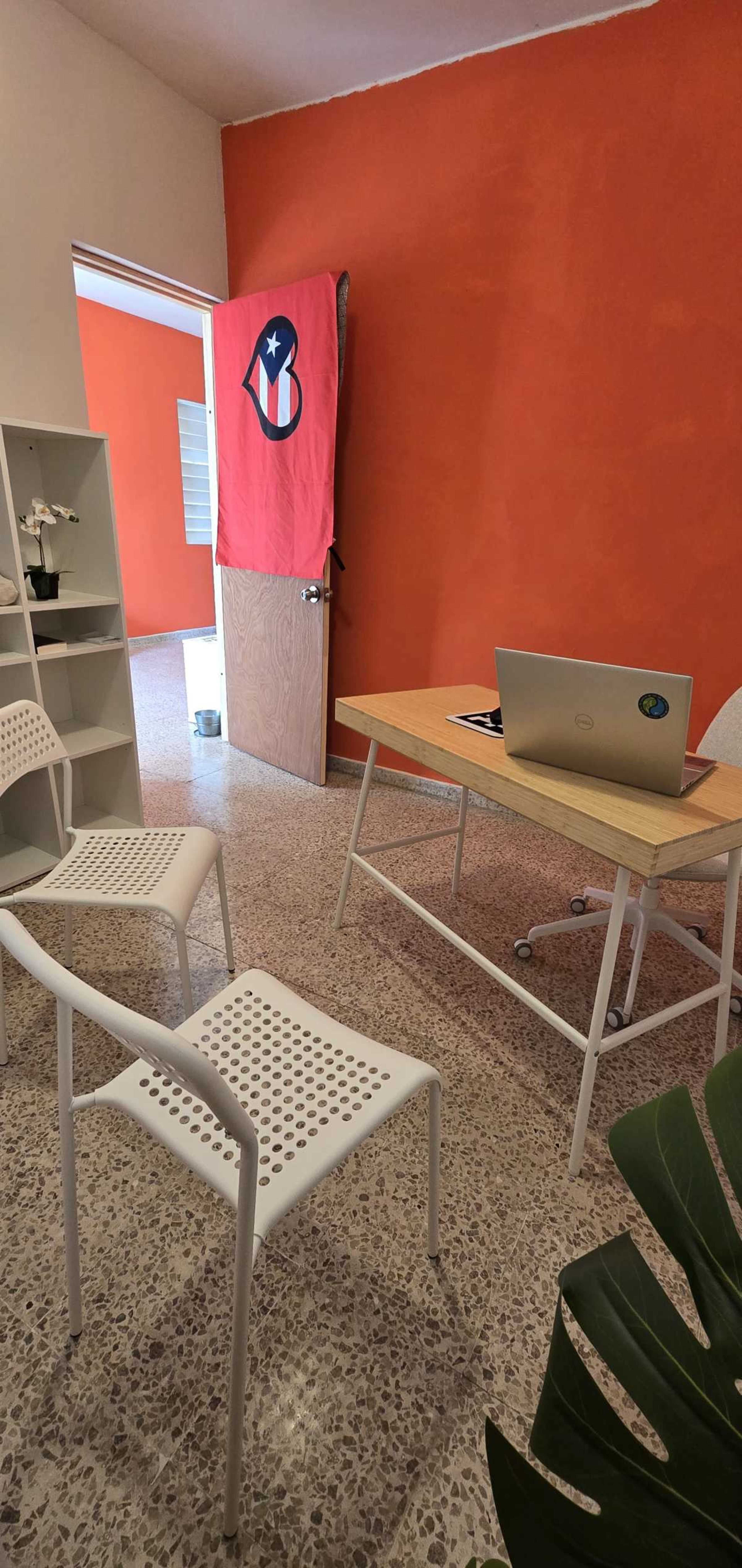 The image shows a small office space with a wooden desk and a laptop, flanked by two white chairs, and a red wall displaying a flag.