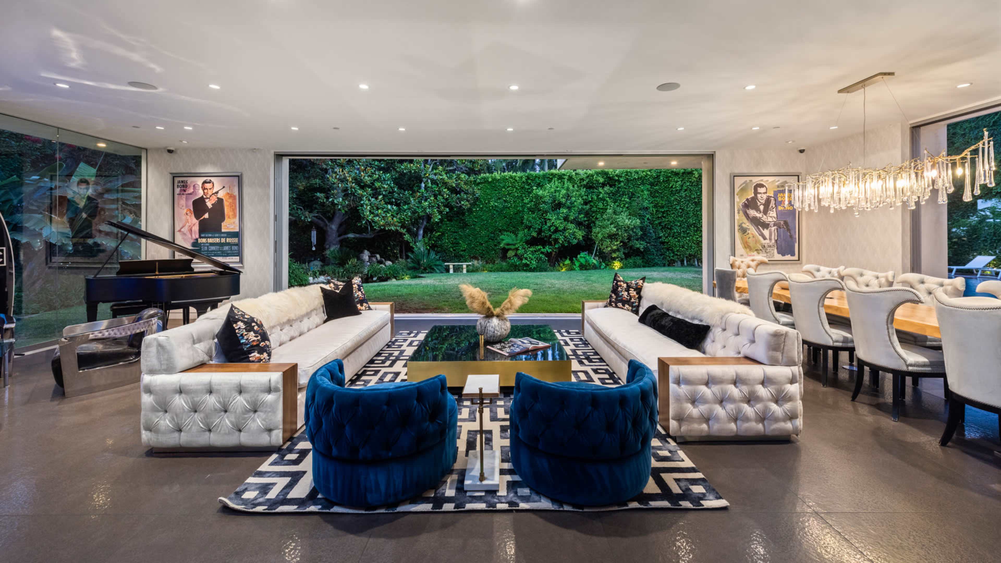 The image shows a modern living room featuring two tufted white sofas, blue accent chairs, a coffee table, and a grand piano, with large windows overlooking a green outdoor space.