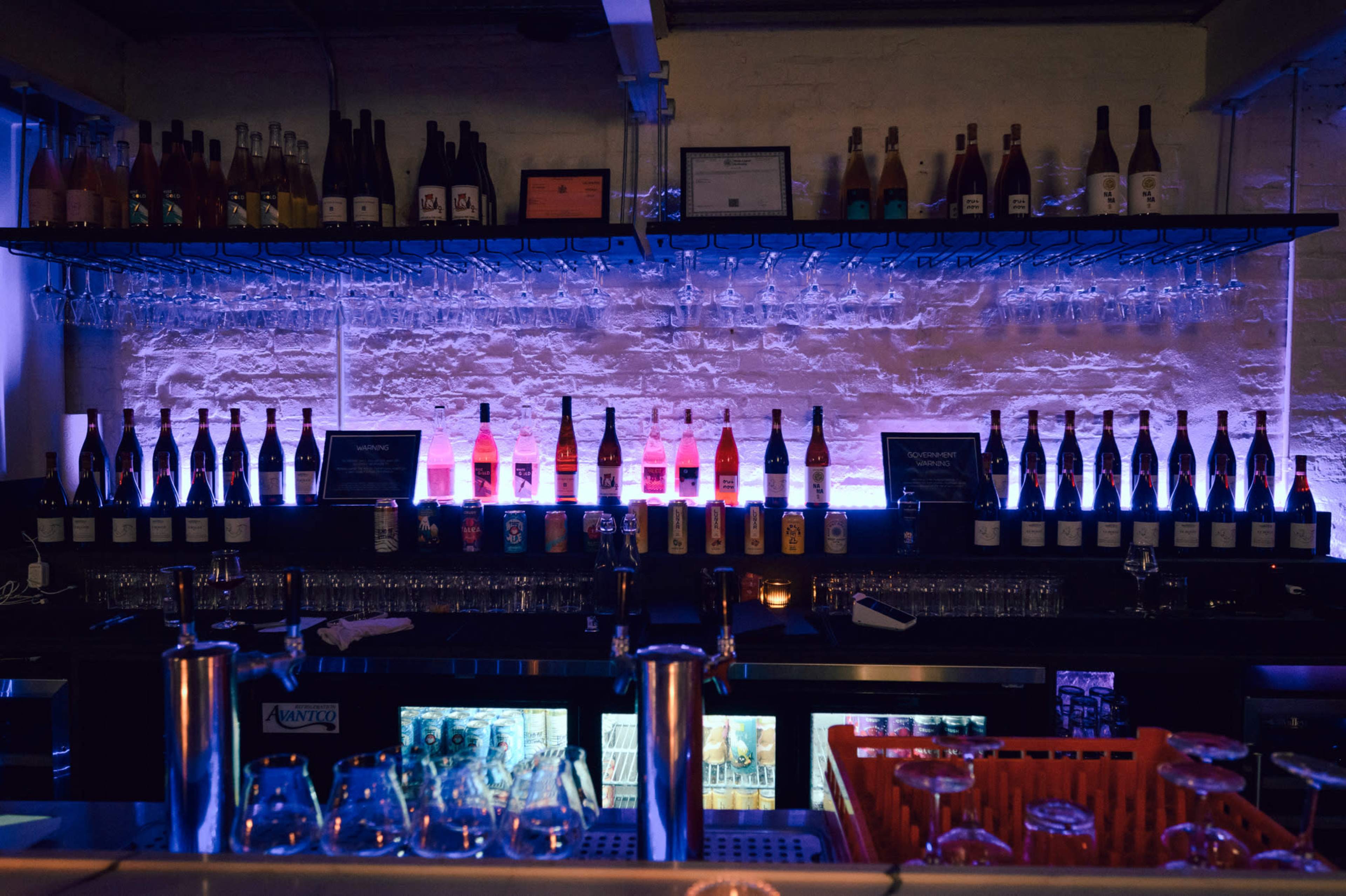A dimly lit bar with a backlit shelf displaying various bottles and glasses.