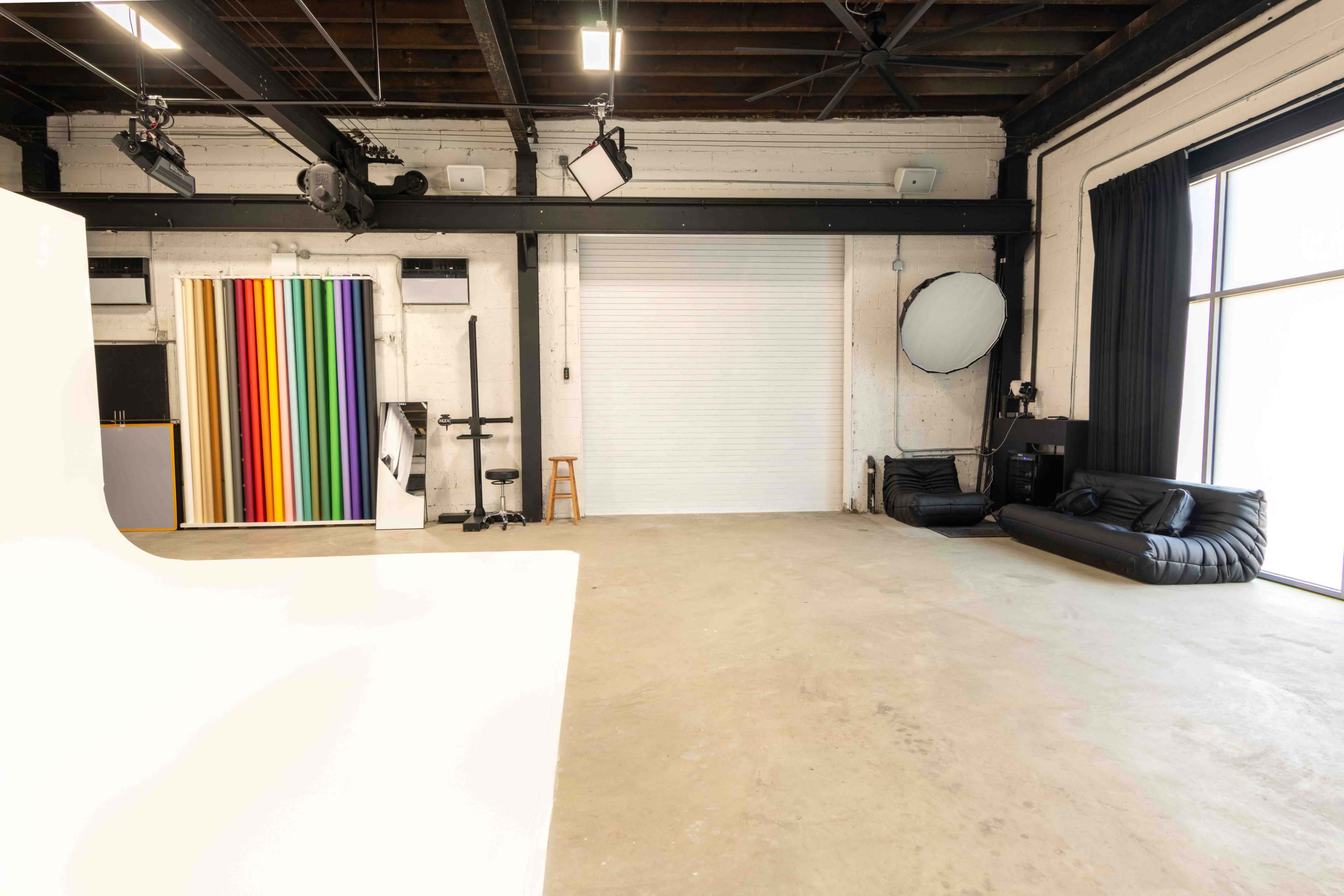 The image shows a spacious photography studio featuring a colorful backdrop, a couch, and equipment setup against a plain concrete floor.