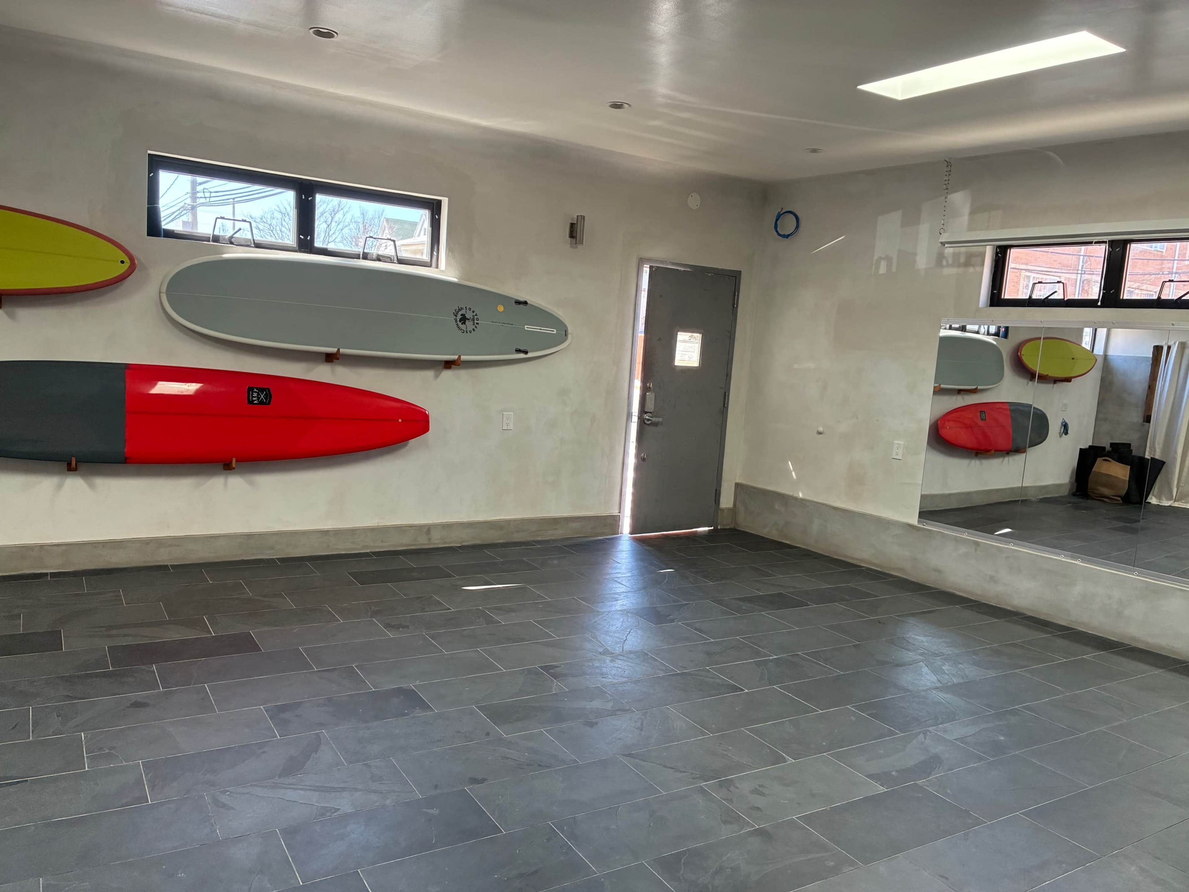 The image shows a room with grey tiled flooring and surfboards mounted on the walls, along with a large mirror and a door leading outside.