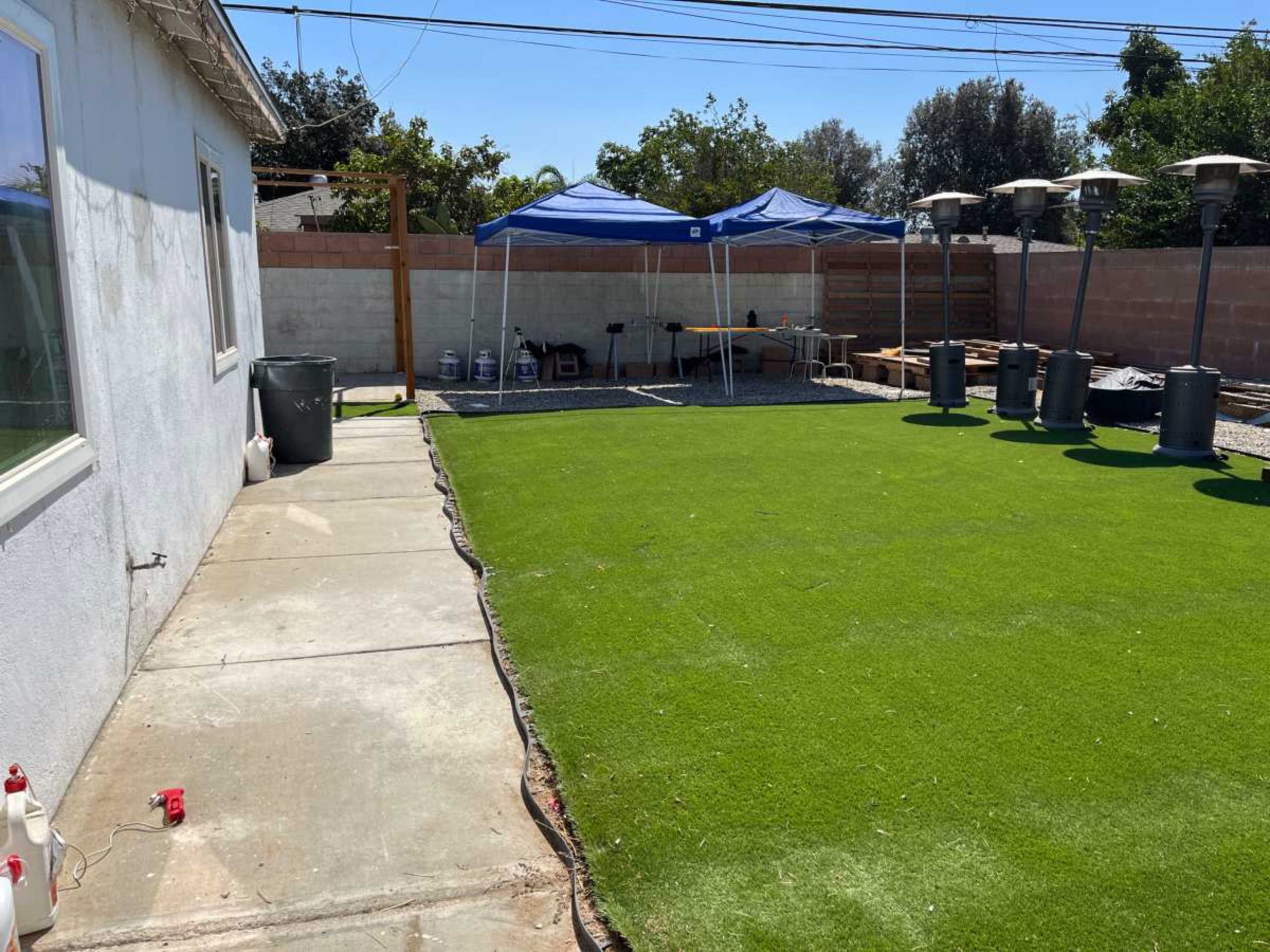 The image shows a backyard with a green artificial lawn, two blue tents set up, and several patio heaters along a fence.