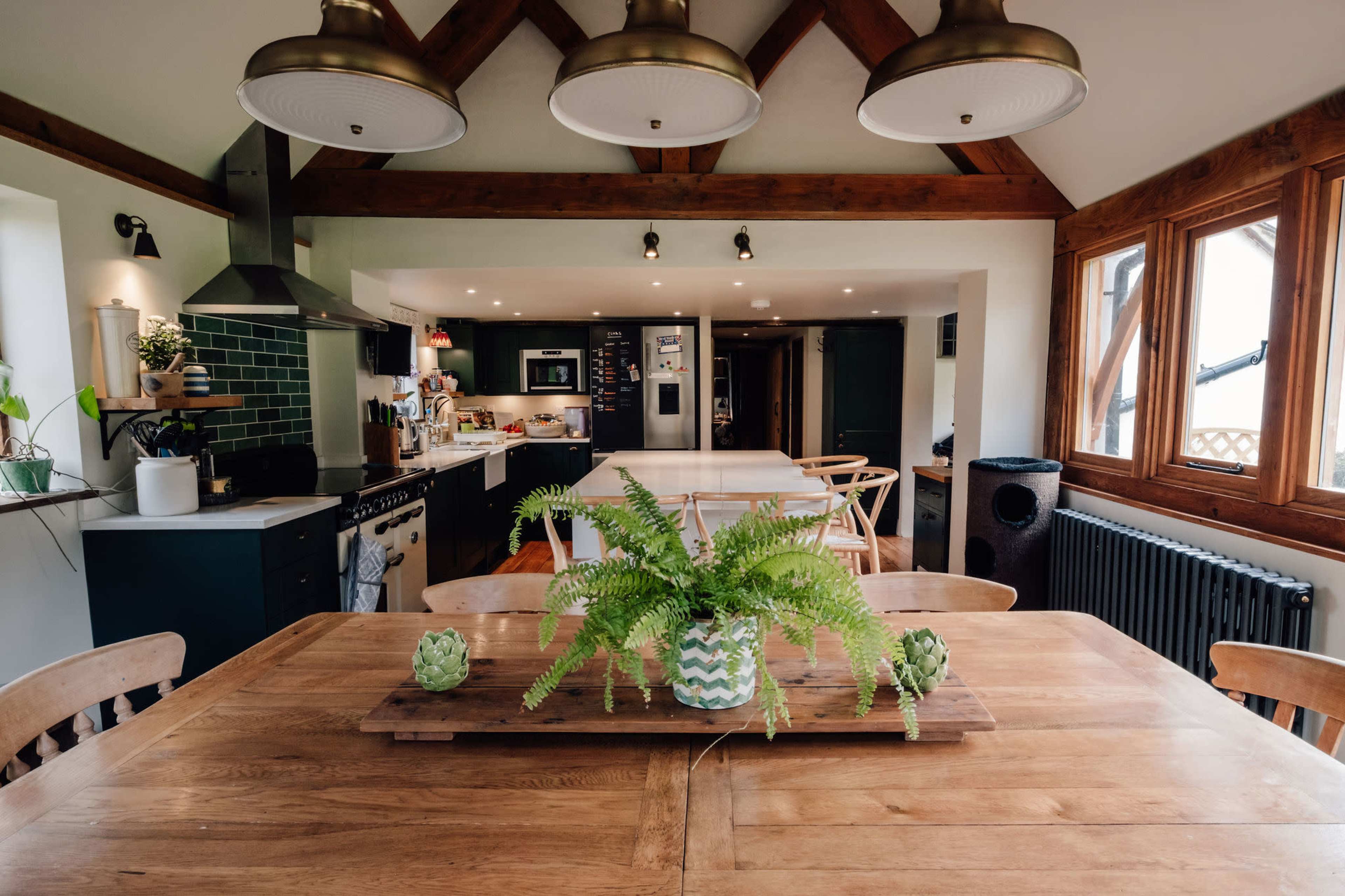 The image shows a modern kitchen and dining area with wooden beams, a large table at the center, and various kitchen appliances in the background.