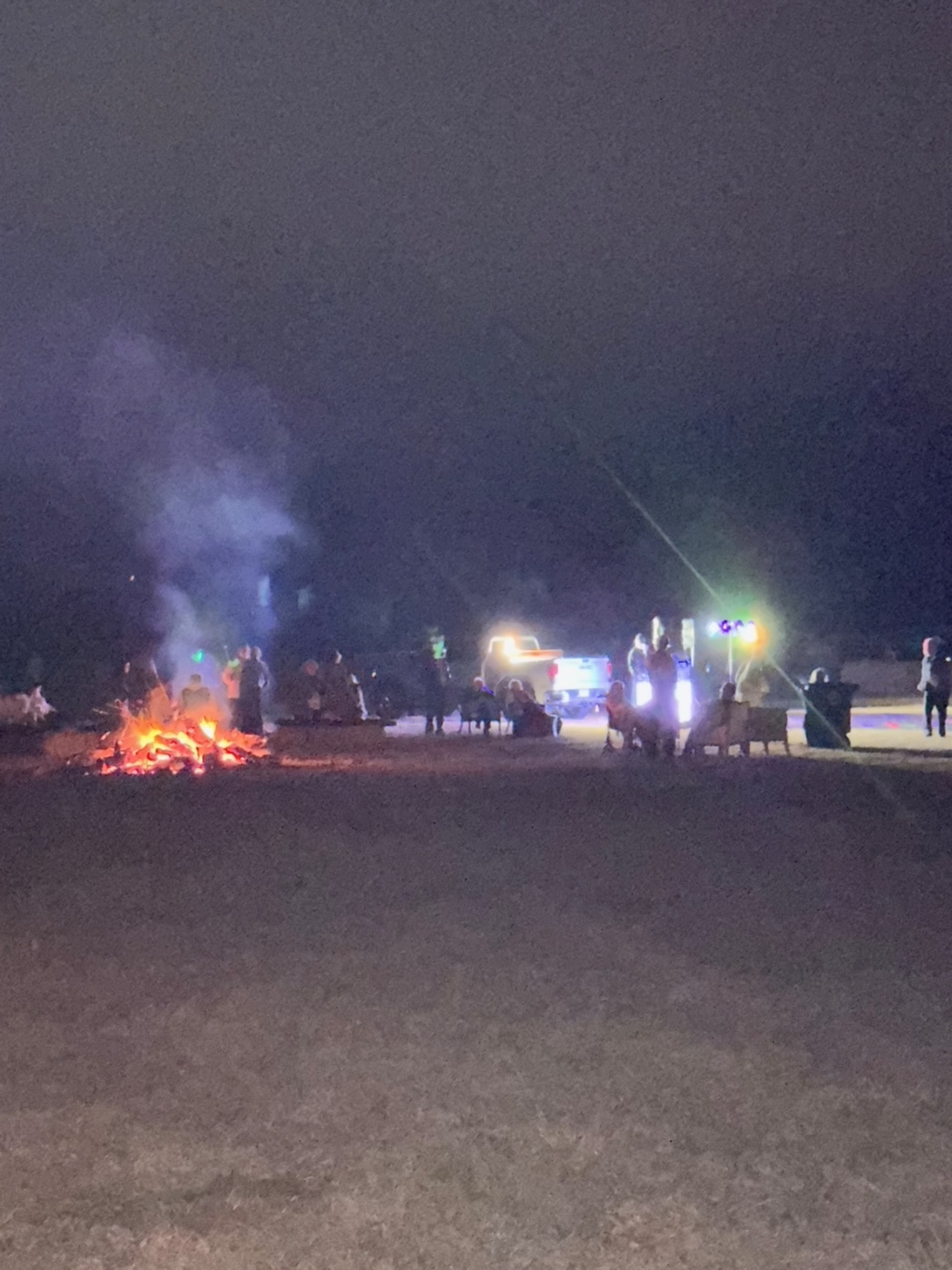 A group of people gathers around a fire at night, while vehicles with headlights illuminate the scene.