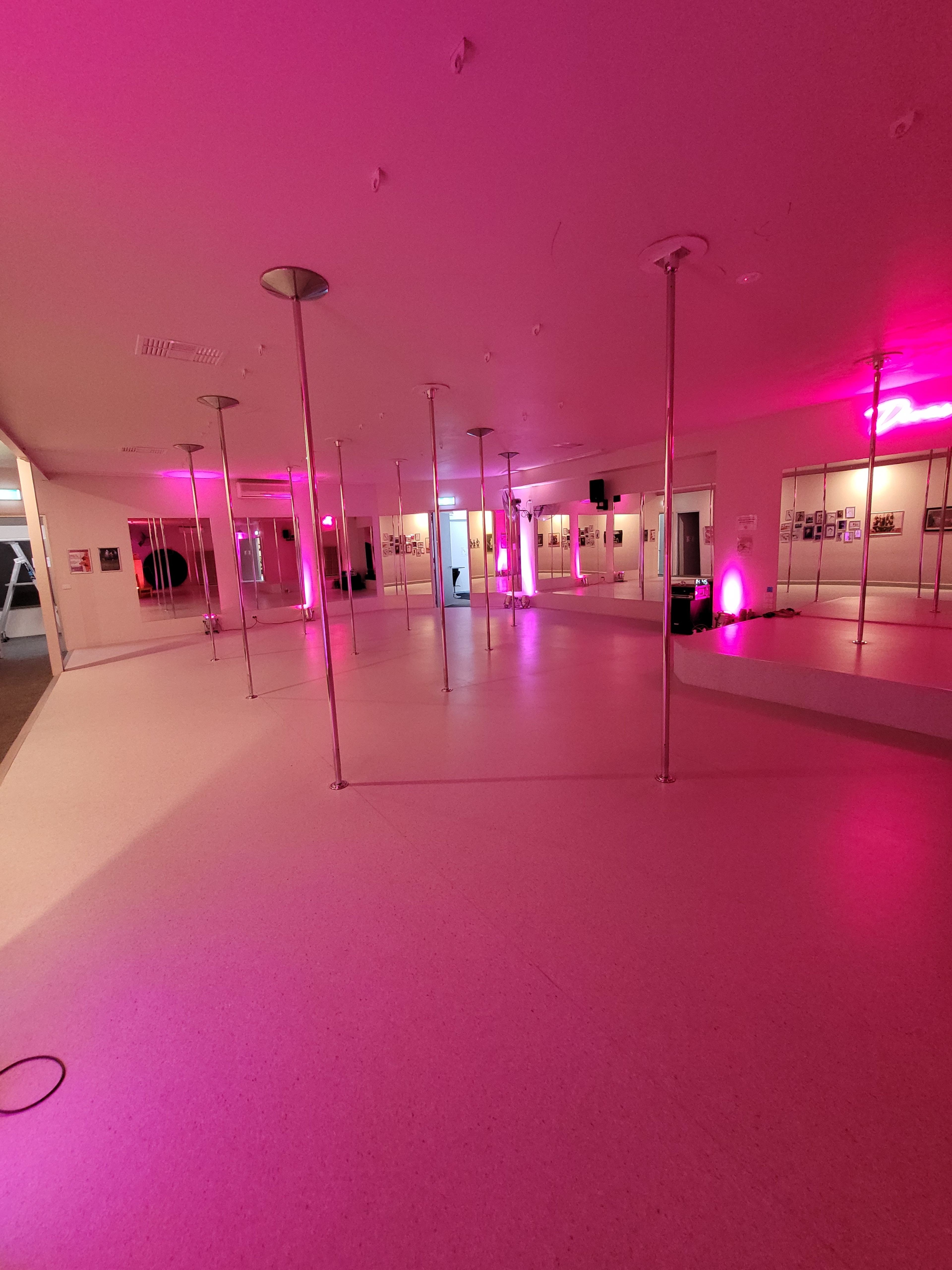 A spacious dance studio with multiple poles and soft pink lighting.