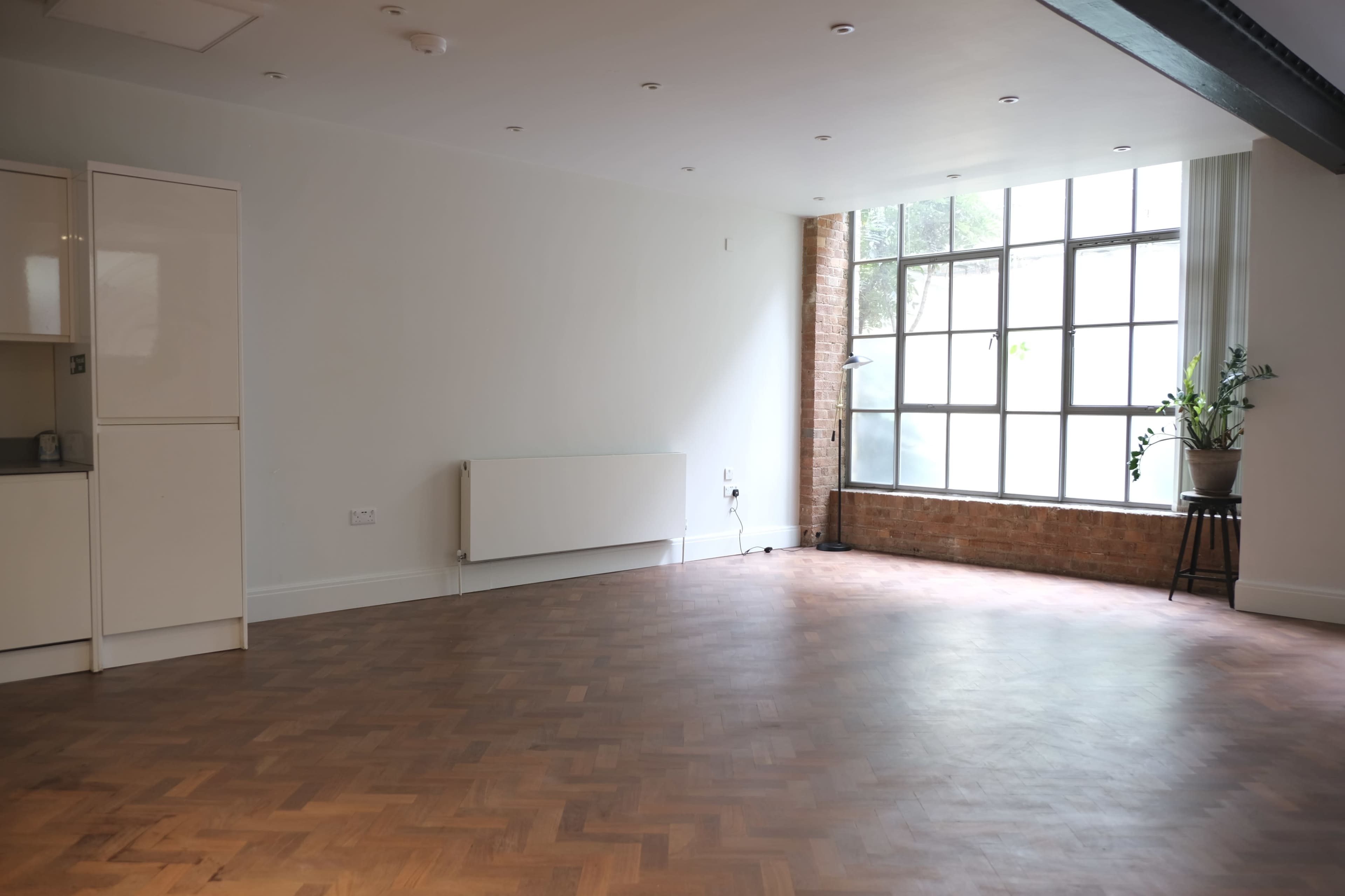 A spacious, empty room with wooden herringbone flooring, large windows, and a plant in the corner.