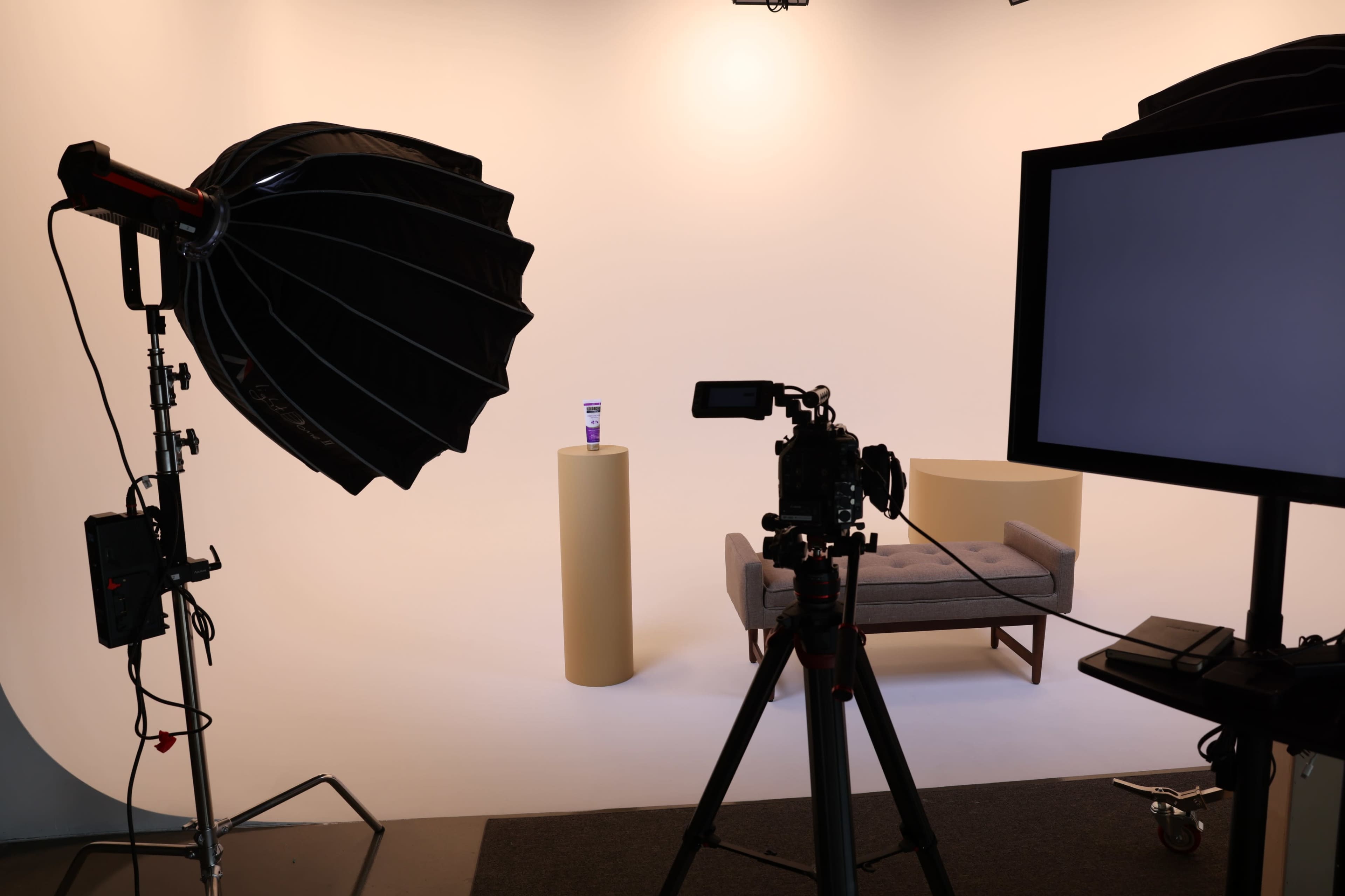 A photography studio setup with a large softbox light, a camera on a tripod, and a pedestal displaying a product in front of a minimalist backdrop.