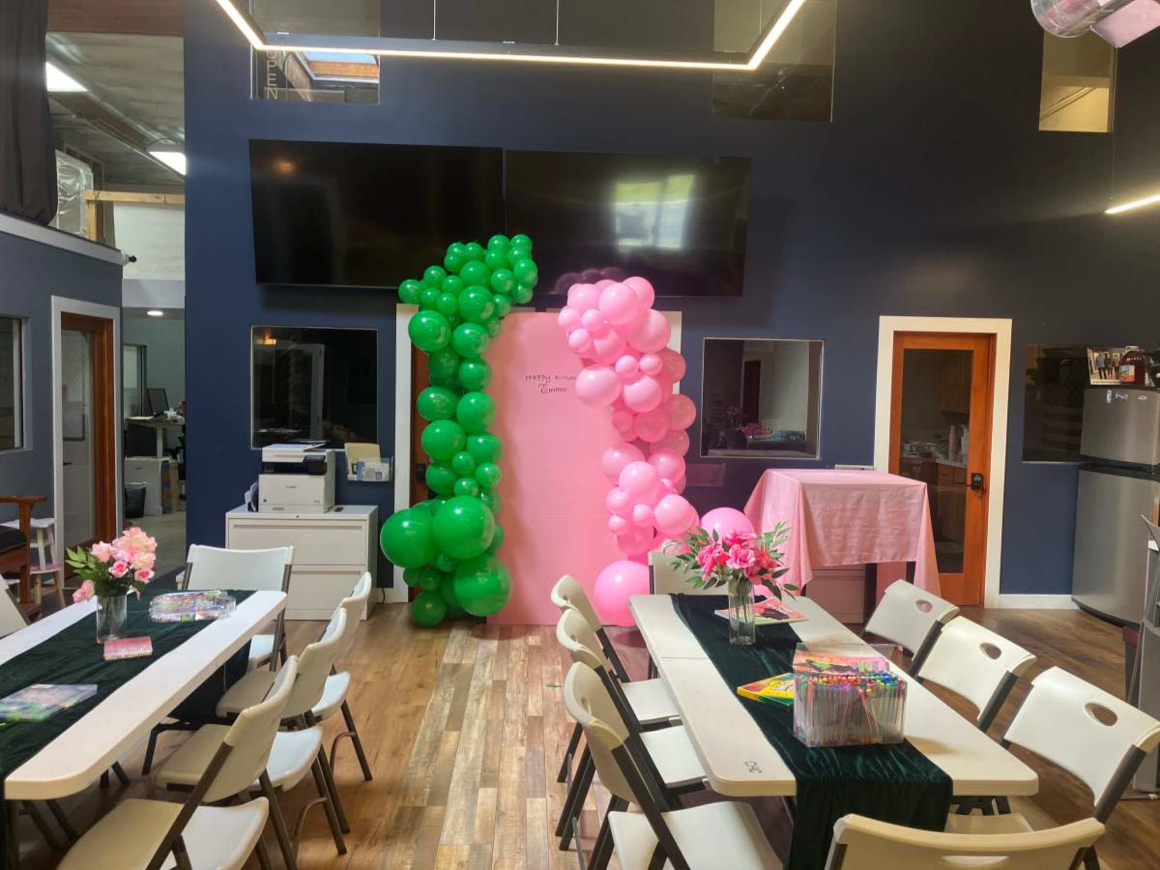 The image shows a decorated indoor event space with a large pink wall adorned with green and pink balloons, tables set with flowers, and artwork supplies arranged.