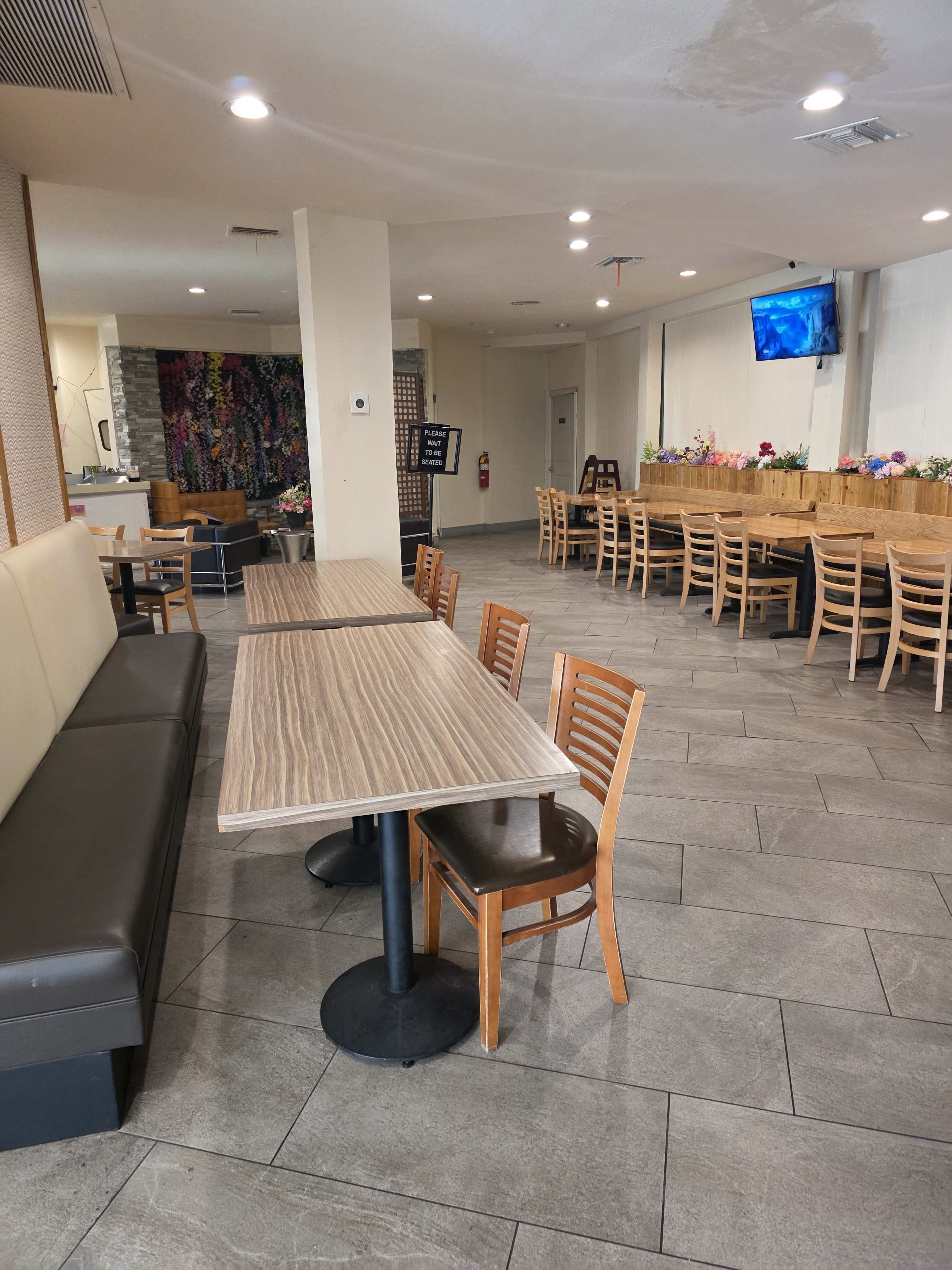 The image shows an empty restaurant interior with wooden tables and chairs, a large booth along one wall, and a television mounted on the opposite wall.