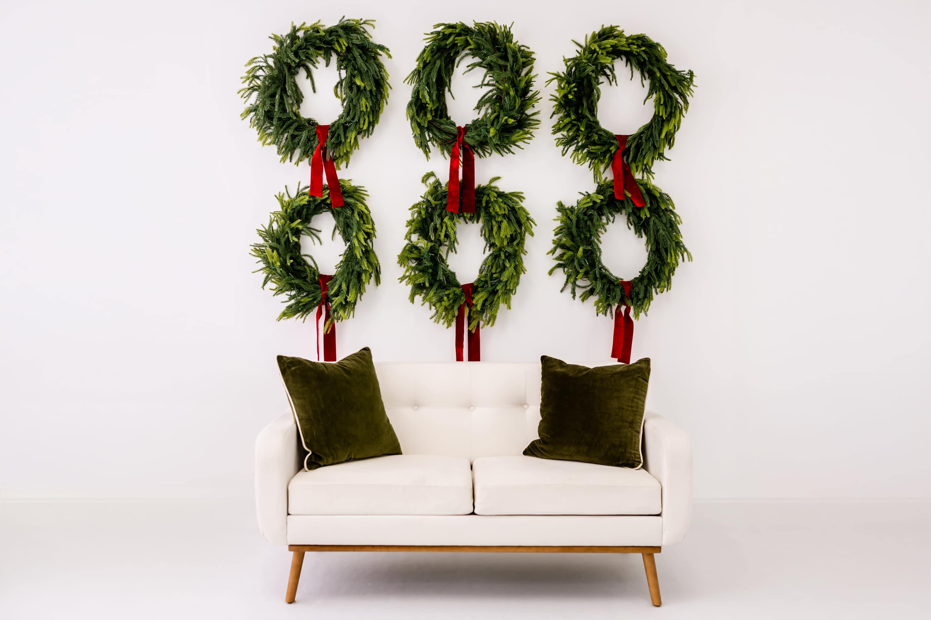 A white couch with green cushions is placed in front of a wall adorned with six hanging wreaths tied with red ribbons.