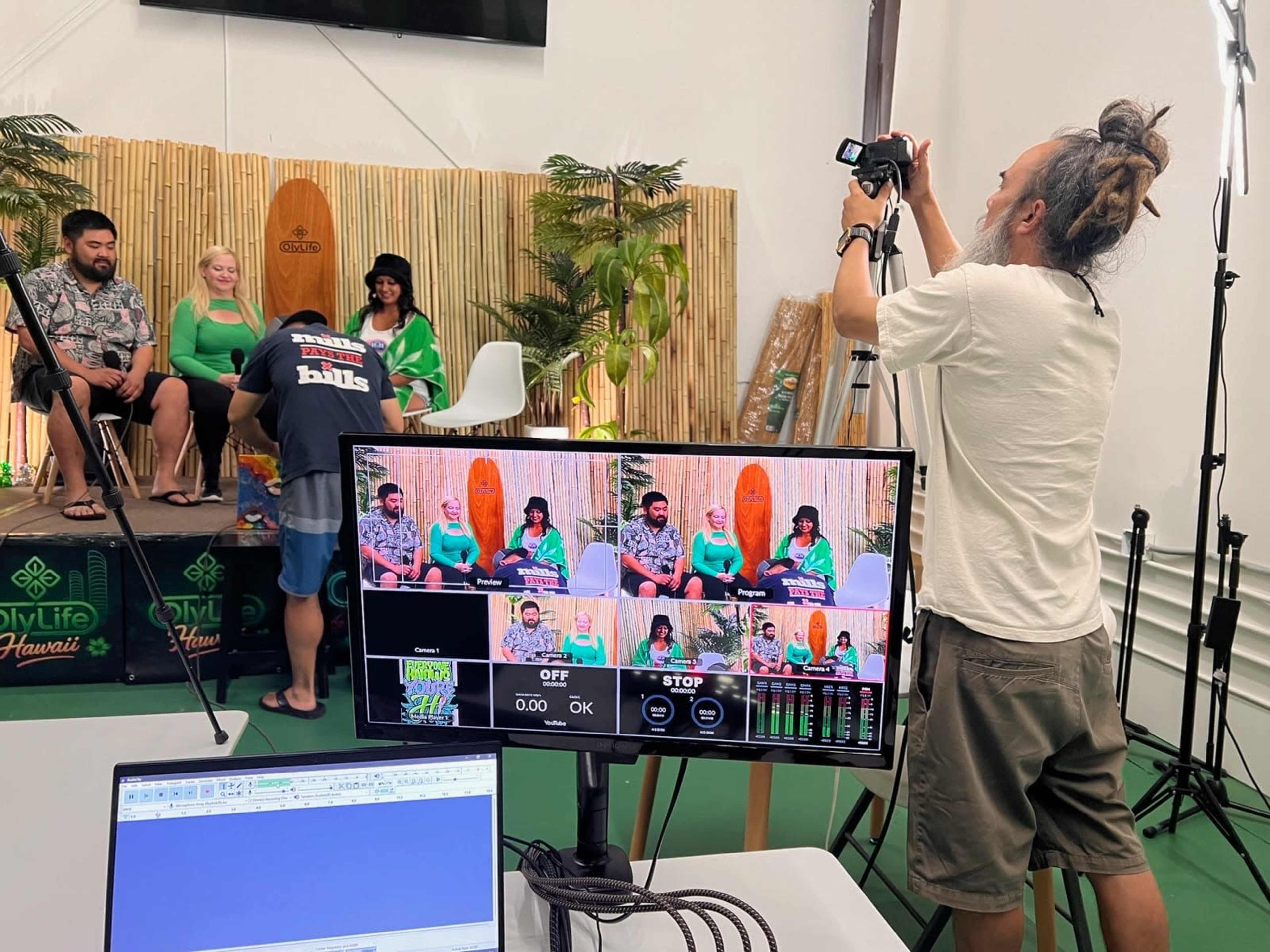 A person is operating a camera while four individuals sit on a stage decorated with tropical plants and surfboards, preparing for a shoot in a studio setting.