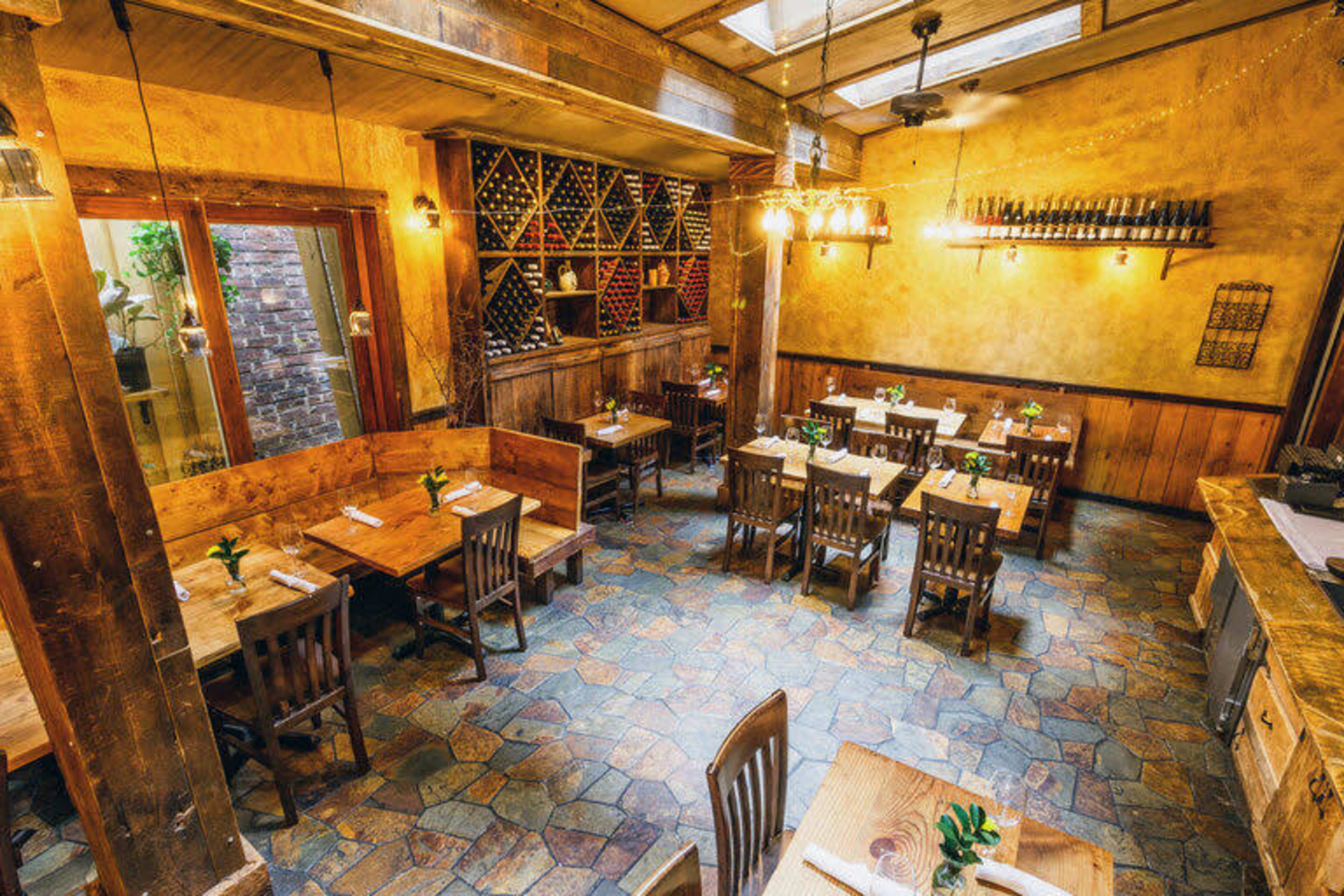 The image shows a rustic restaurant interior with wooden tables, chairs, and a wine rack on the wall.