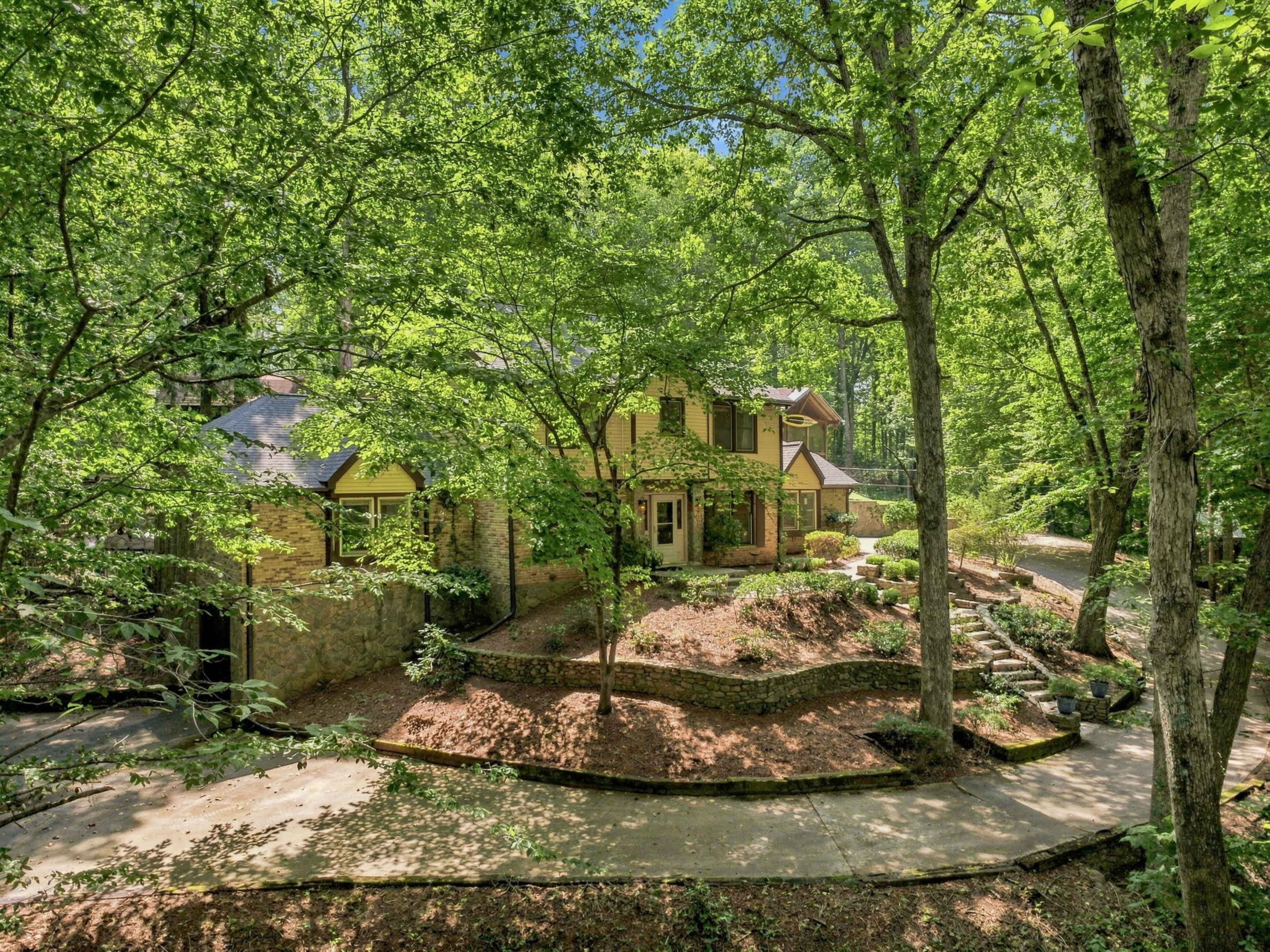 A two-story brick house is surrounded by dense greenery, with a winding driveway leading to its entrance.