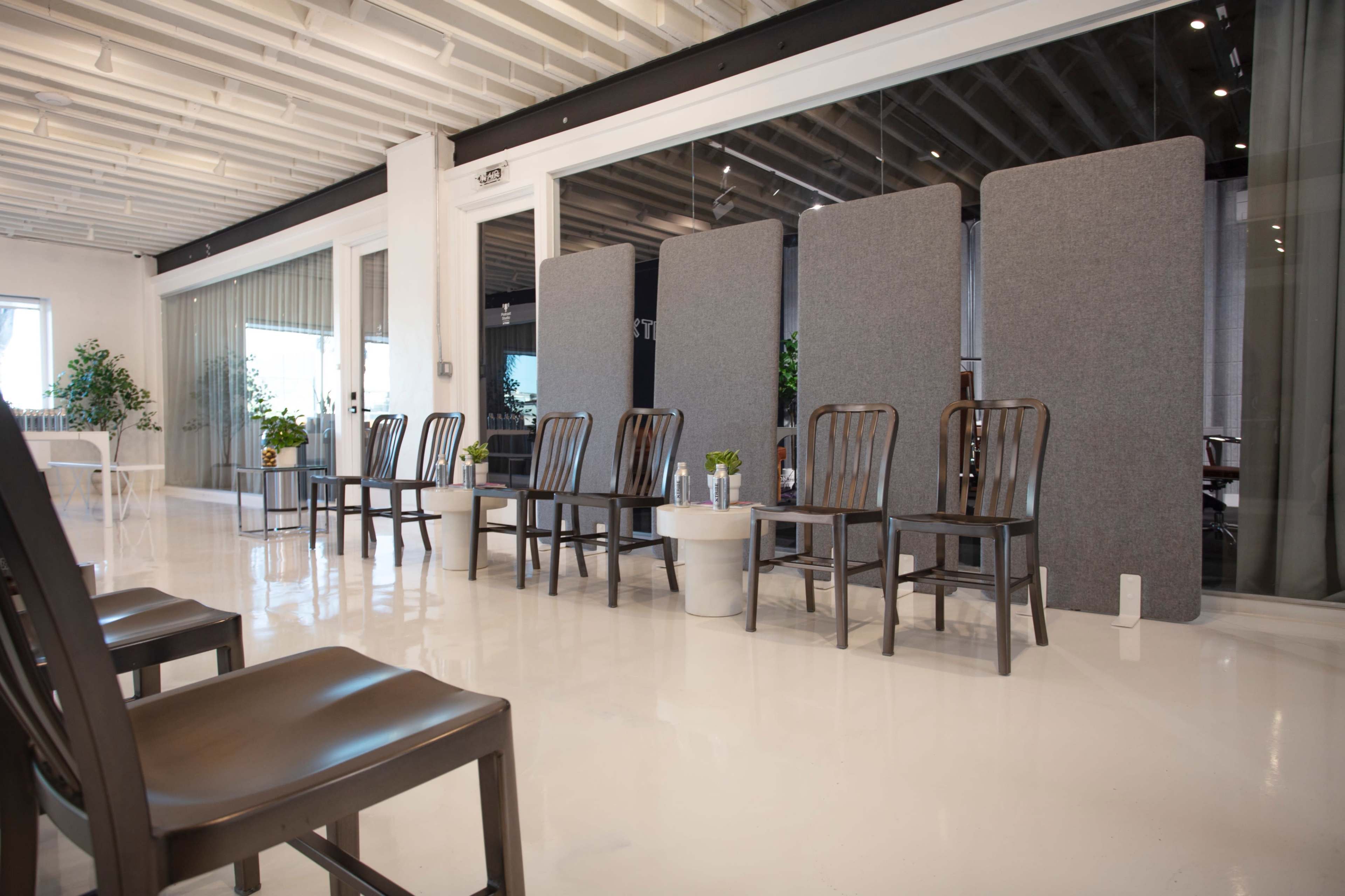 A row of wooden chairs is positioned in front of gray partitions in a spacious, brightly lit office setting.