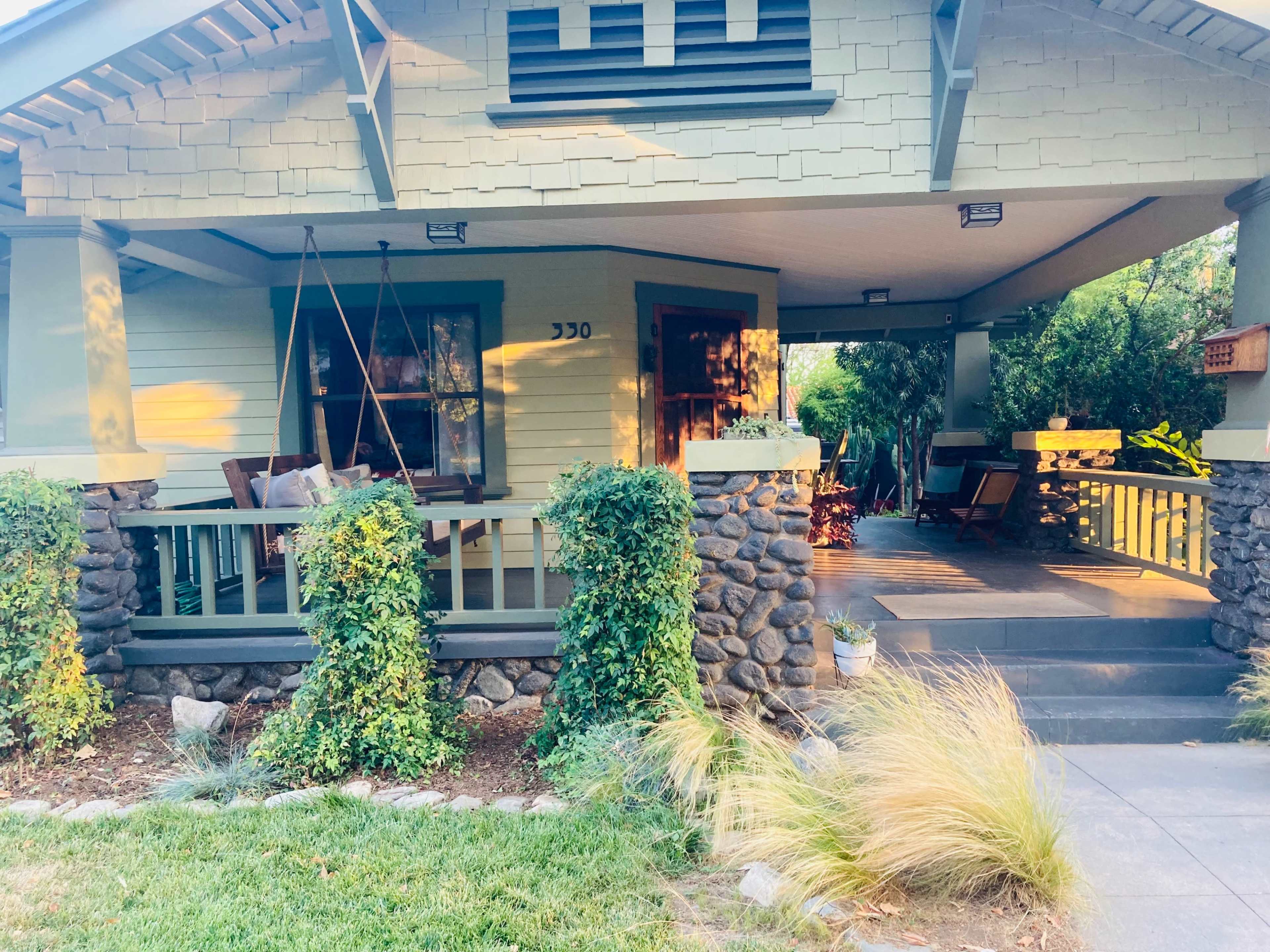 A green craftsman-style house with a porch, surrounded by stone pillars and lush plants.
