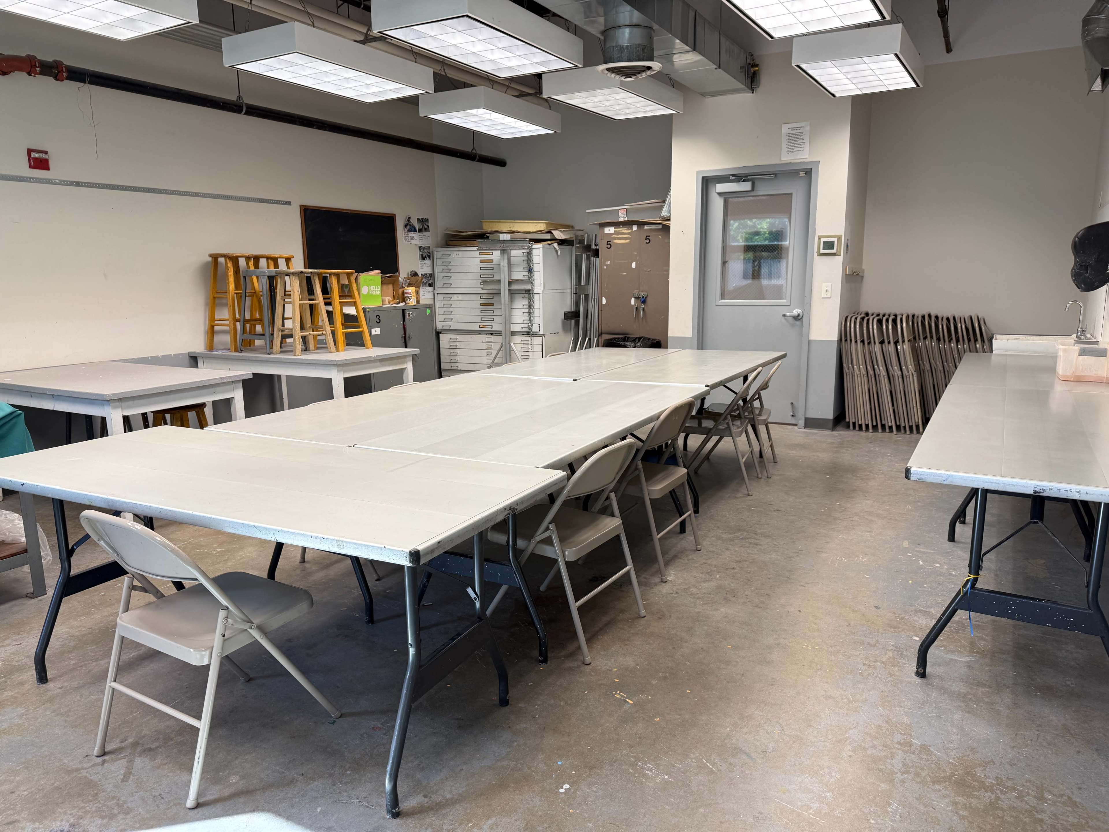 The image shows a spacious workshop or studio with several folding tables arranged in the center and various workstations along the walls, illuminated by overhead lights.