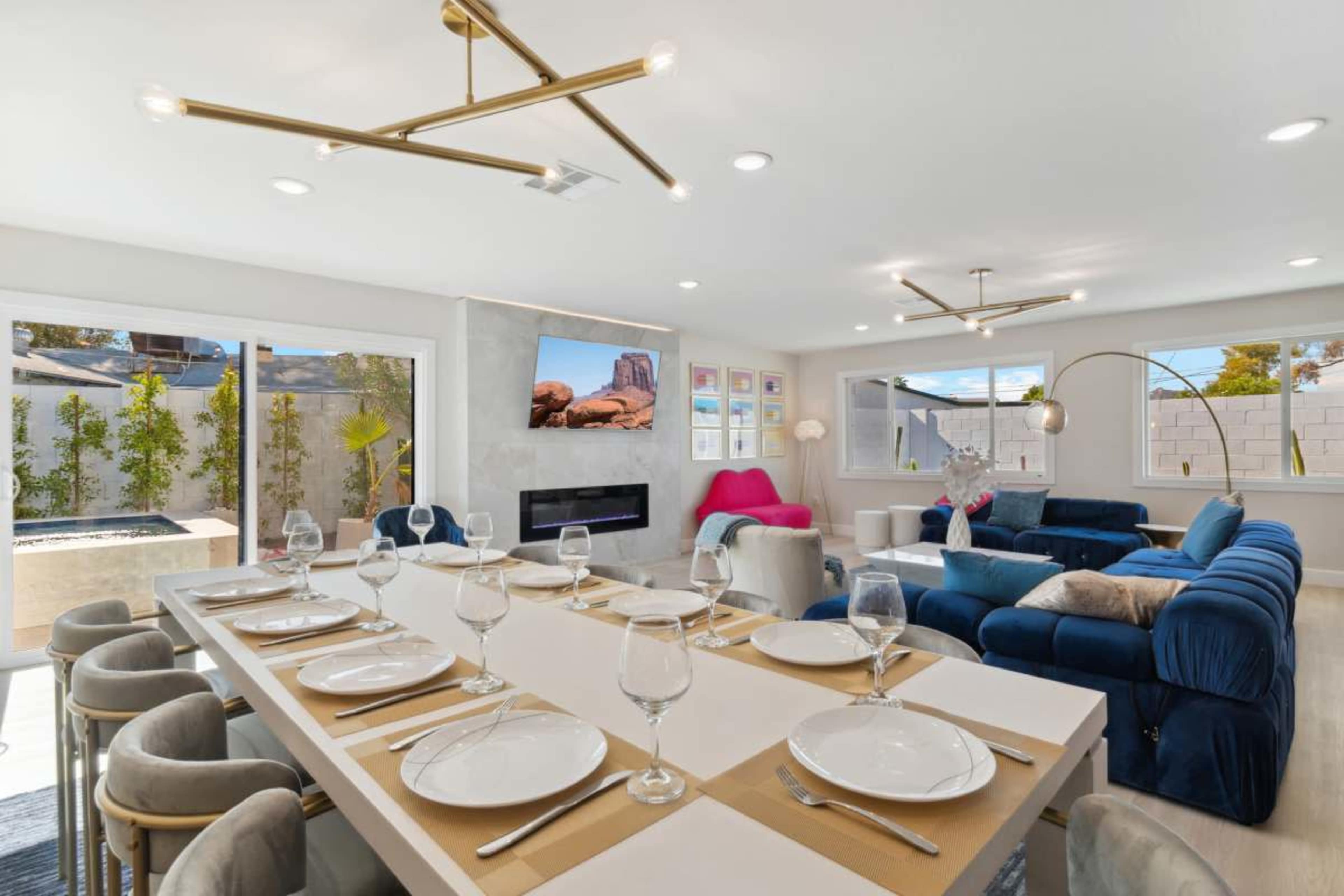 The image shows a modern, well-lit living and dining area with a large dining table set for eight, a sectional sofa, and a wall-mounted television.