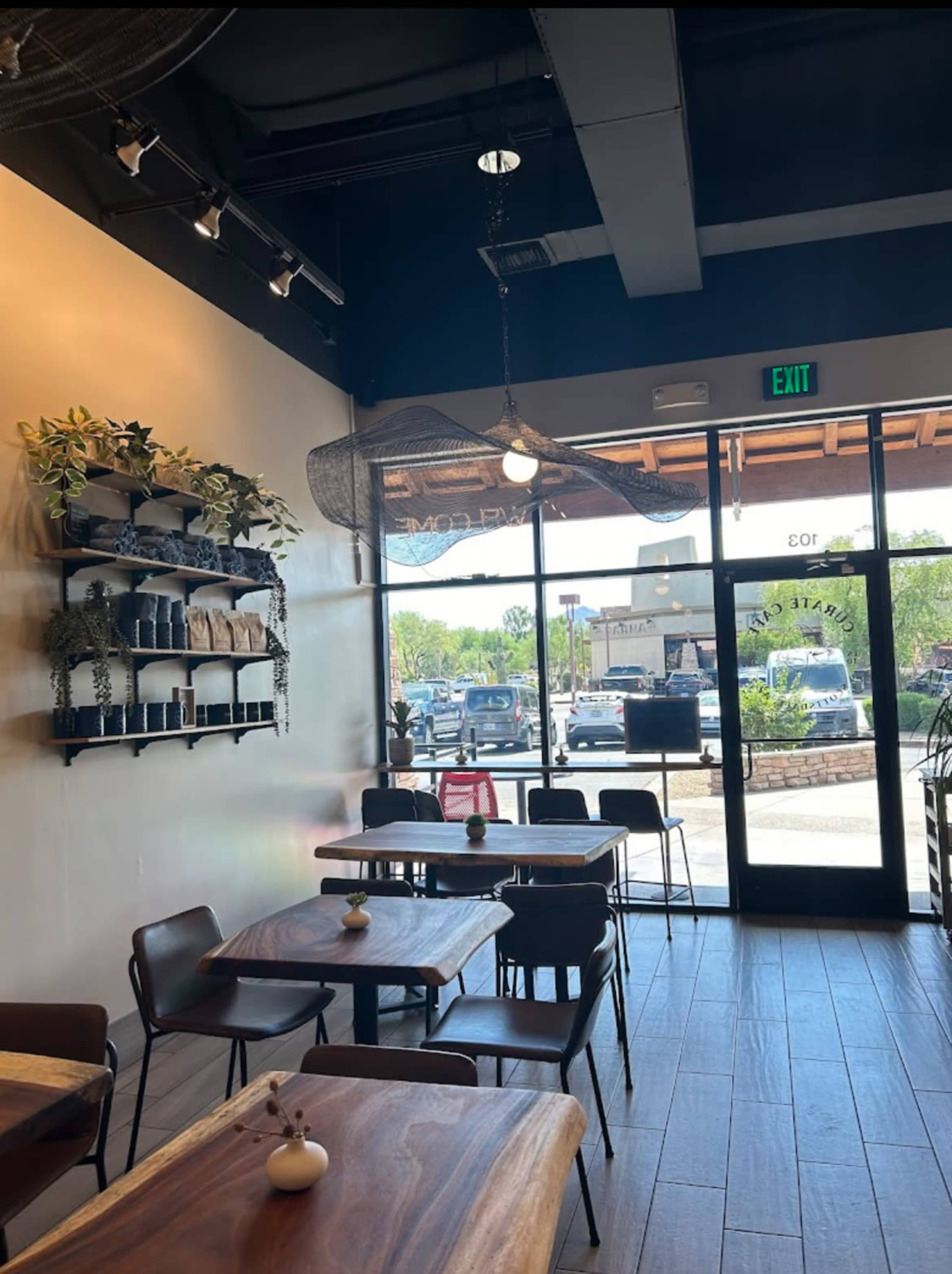 The interior of a café featuring wooden tables, chairs, and plants on a wall shelf, with large windows displaying a view of parked cars outside.
