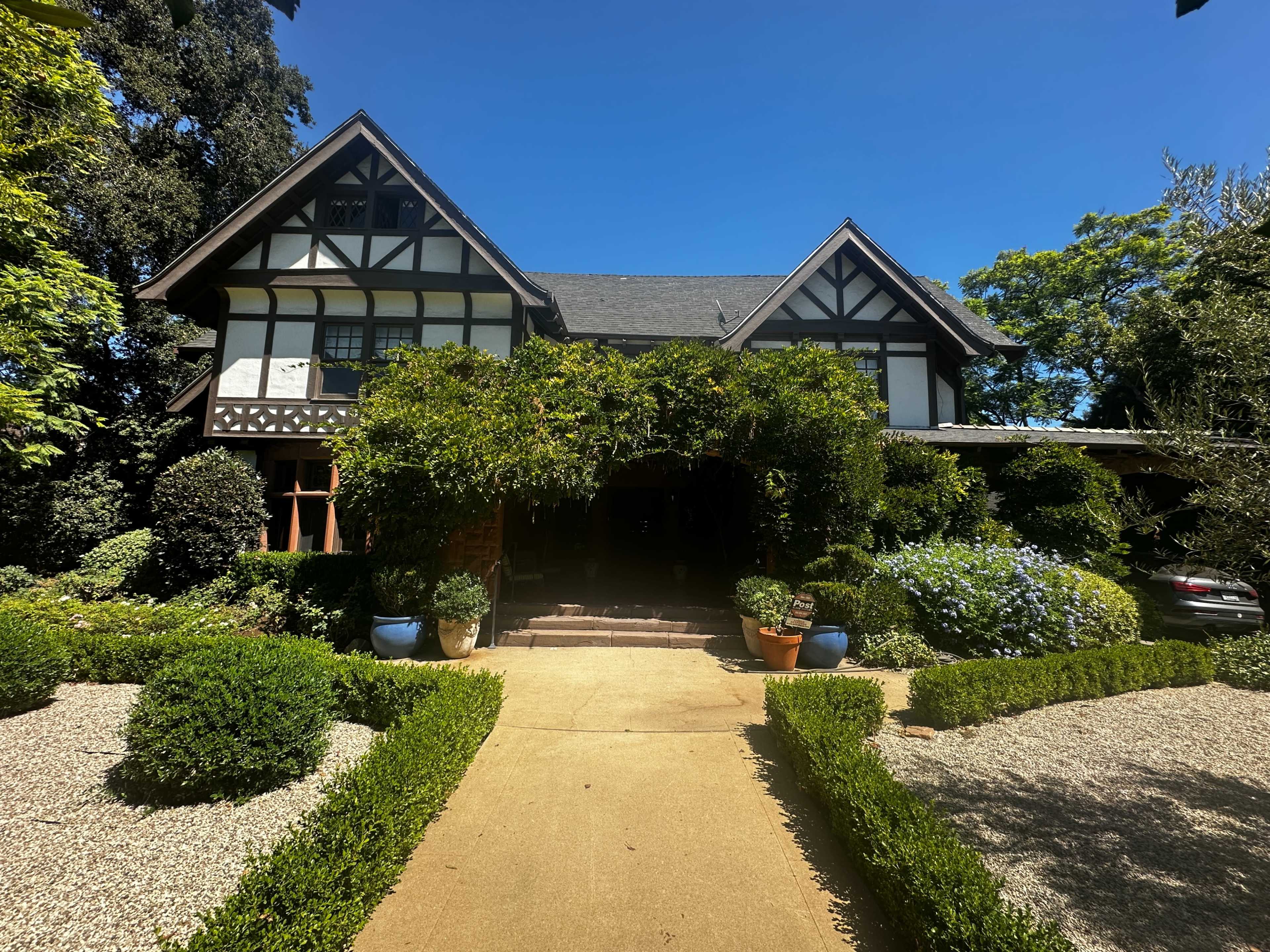 A large, Tudor-style house with a prominent entrance is surrounded by lush greenery and well-manicured landscaping.